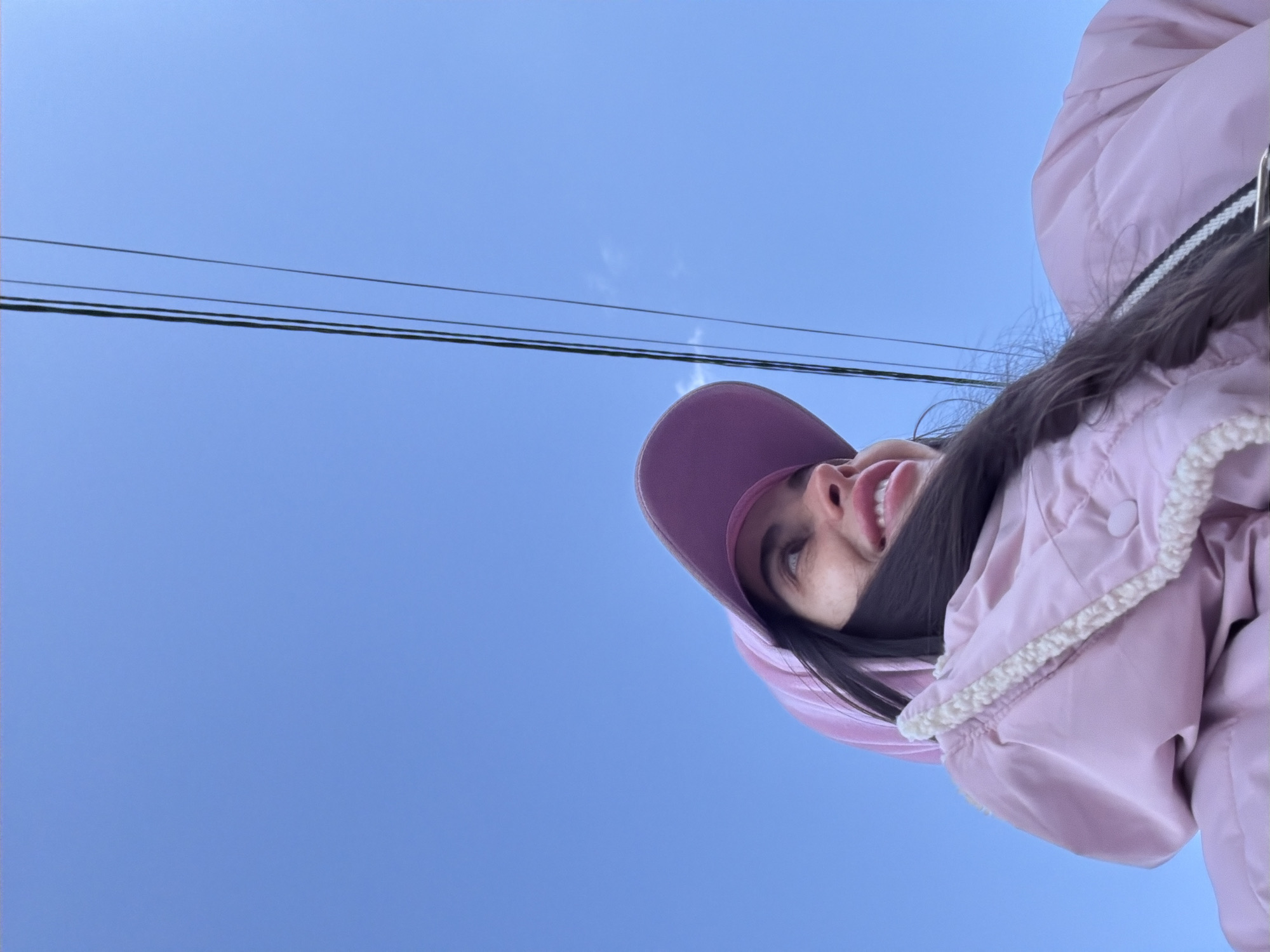 A female subject is depicted from a low-angle perspective, observed from the chest up, looking upward and to the right with a visible smile. She is wearing a light pink padded jacket accented with cream-colored fleece trim, a pink baseball cap, and a pink hoodie beneath the jacket. Her long dark hair is partially visible. A black and white striped strap is present over her right shoulder. The background consists of a clear blue sky with three dark utility lines extending diagonally from the upper left portion of the frame towards the lower right. A single small, wispy white cloud is visible in the upper right quadrant of the sky. The scene is located in the city of Saint Petersburg, Russia.