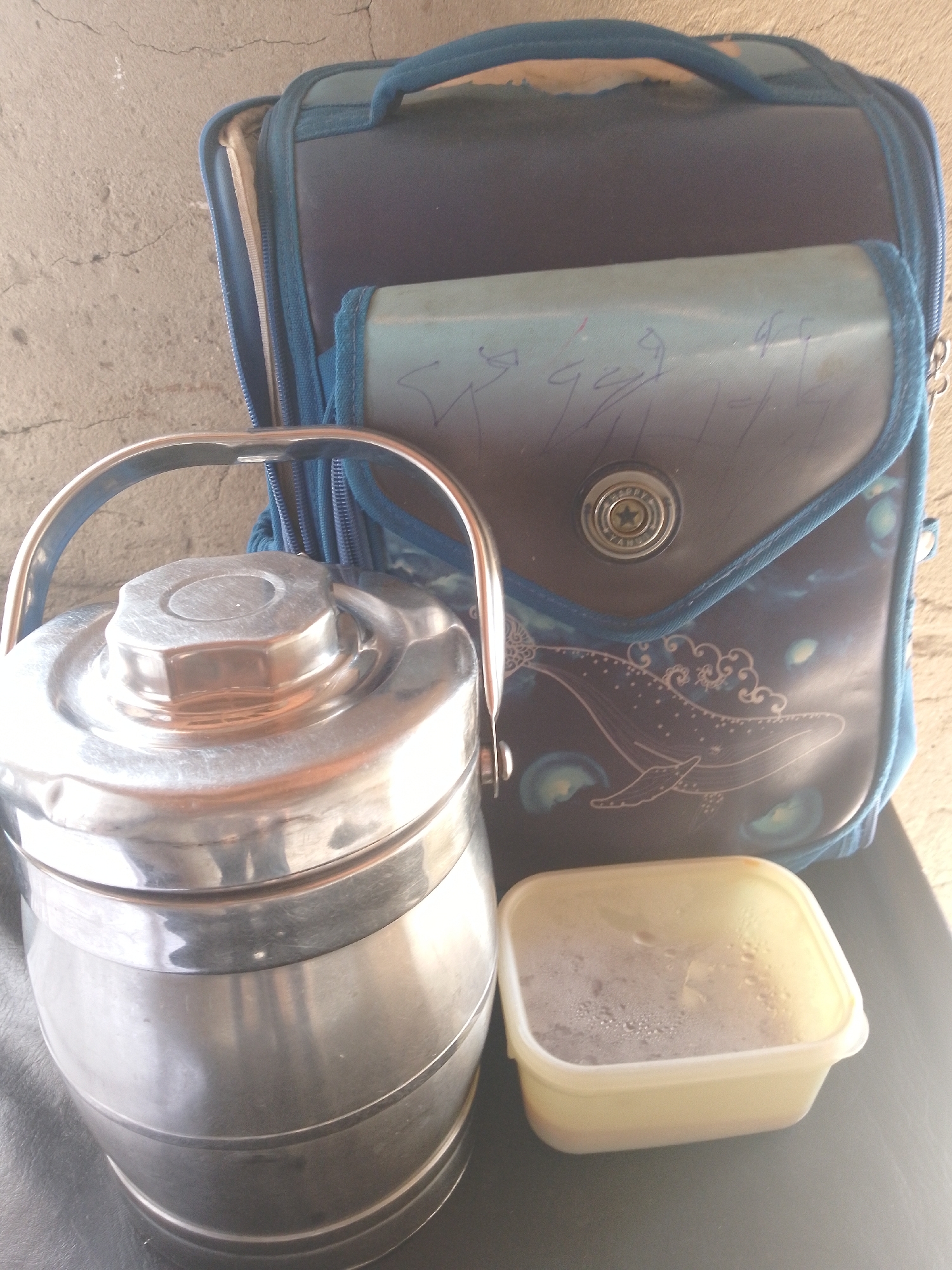 The image depicts a close-up, overhead view of lunch items placed on a dark surface, likely a table or ground, with a textured, light-colored wall in the background. To the left, a metallic, multi-tiered tiffin carrier with a rounded lid and a silver handle is prominently featured. Next to it, to the right, is a translucent plastic container filled with a frothy, dark liquid, possibly a beverage or soup. Behind these food containers, a blue backpack with a flap is visible. The backpack has a white drawing of a whale and some stylized clouds on its front, and a circular emblem with a star is affixed to the flap. The backpack also features blue trim and zippers. The overall scene appears to be an outdoor setting, with the sunlight casting distinct shadows and highlighting the metallic sheen of the tiffin carrier. The environment's rough, concrete-like wall suggests a possibly urban or semi-urban location. Based on the visual cues, the image is likely from Serti, Nigeria, given the context provided.