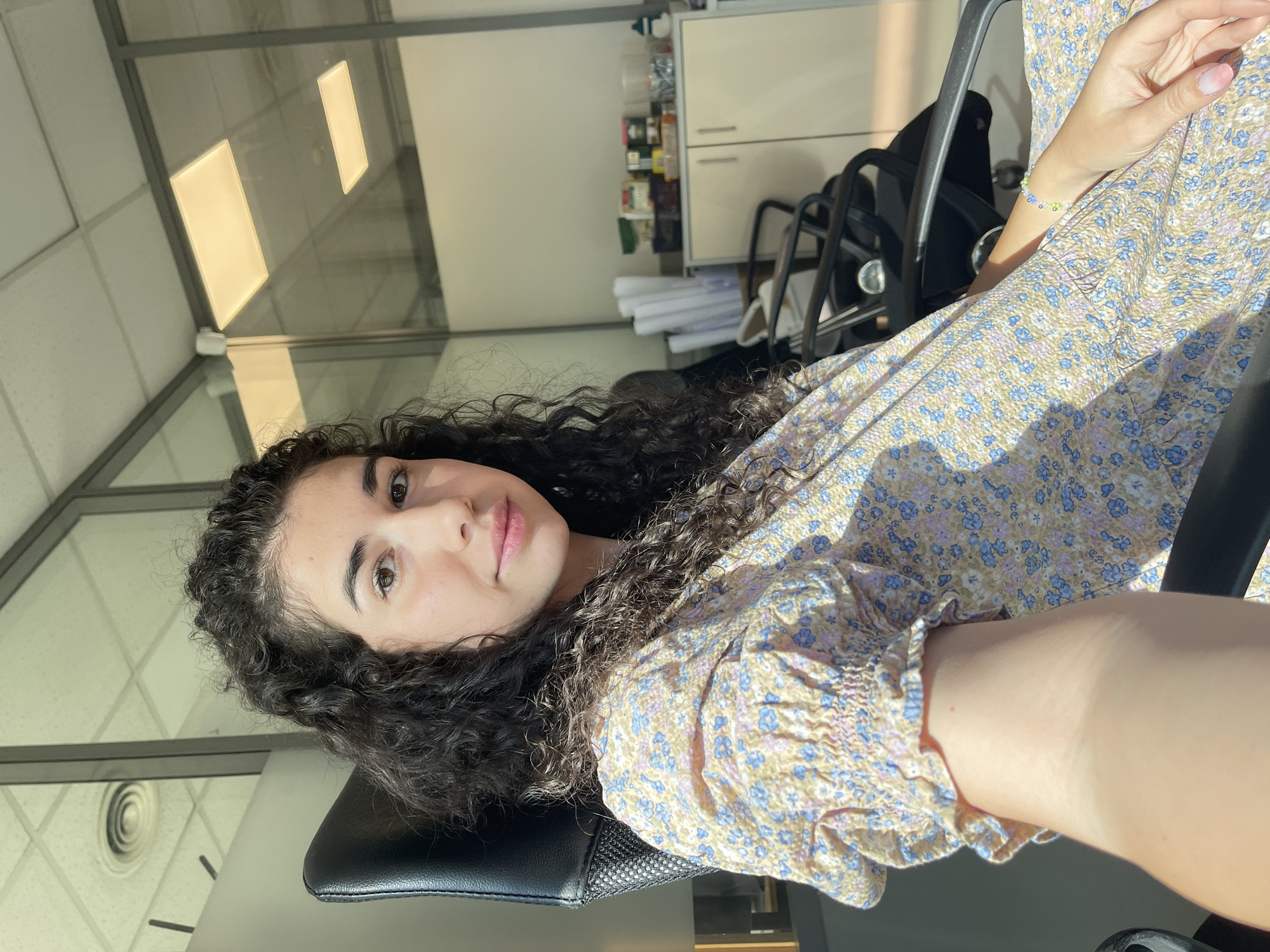 A young woman with dark, curly hair is sitting in an office chair, facing the camera. She is wearing a light-colored, floral-print dress. Her left arm is extended towards the camera, and she is holding a smartphone to take a selfie. The background appears to be an office with glass partitions, fluorescent lighting, and office furniture, including a cabinet with various items on top. The scene is illuminated by natural sunlight streaming in from the right, casting shadows across the room and the woman. The office location appears to be in Sofia, Bulgaria, indicated by the presence of a specific type of air vent seen in the ceiling reflection.