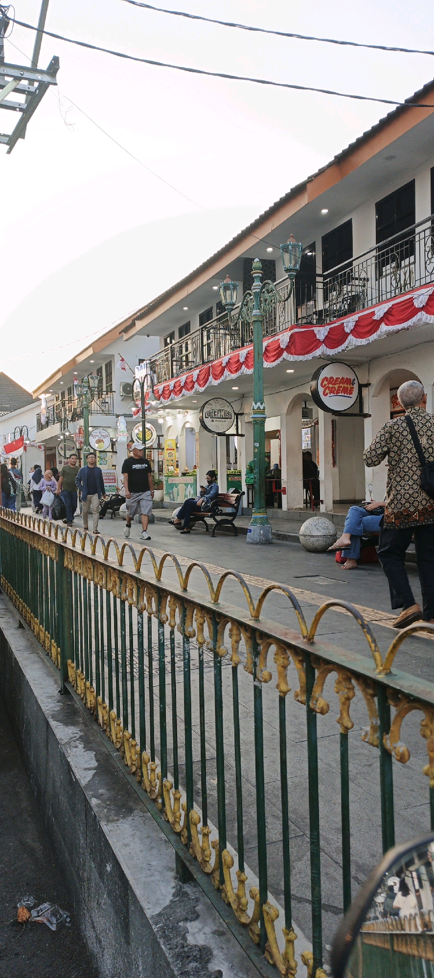 This is a vertical, eye-level, outdoor, medium shot captured during the day in Yogyakarta, Indonesia, likely at Malioboro Street given the architectural style and atmosphere.
The scene depicts a pedestrian walkway alongside a row of colonial-style buildings. The buildings have white facades, dark-framed windows, and balconies with ornate black metal railings. Red and white bunting decorates the balconies, suggesting a festive occasion. Several shops with round signage are visible, including one that reads "CREAM & CREME". Traditional street lamps with green ornate metalwork stand along the walkway.
A green metal fence with yellow decorative accents runs along the left side of the image, separating the walkway from a lower level which appears to be a drainage ditch with some litter visible. The walkway is paved with grey tiles.
Several people are present. In the foreground, a person from behind, wearing a patterned shirt and black pants, walks away from the camera. To their left, another person in blue jeans and a grey top sits on a bench with bare feet resting on the ground. To the far left, a group of people are walking along the street, some carrying bags. Other individuals are seated on benches or walking in the middle ground.
The sky is bright and hazy, indicating a sunny day. The overall impression is a bustling urban environment with a blend of historical architecture and contemporary activity.