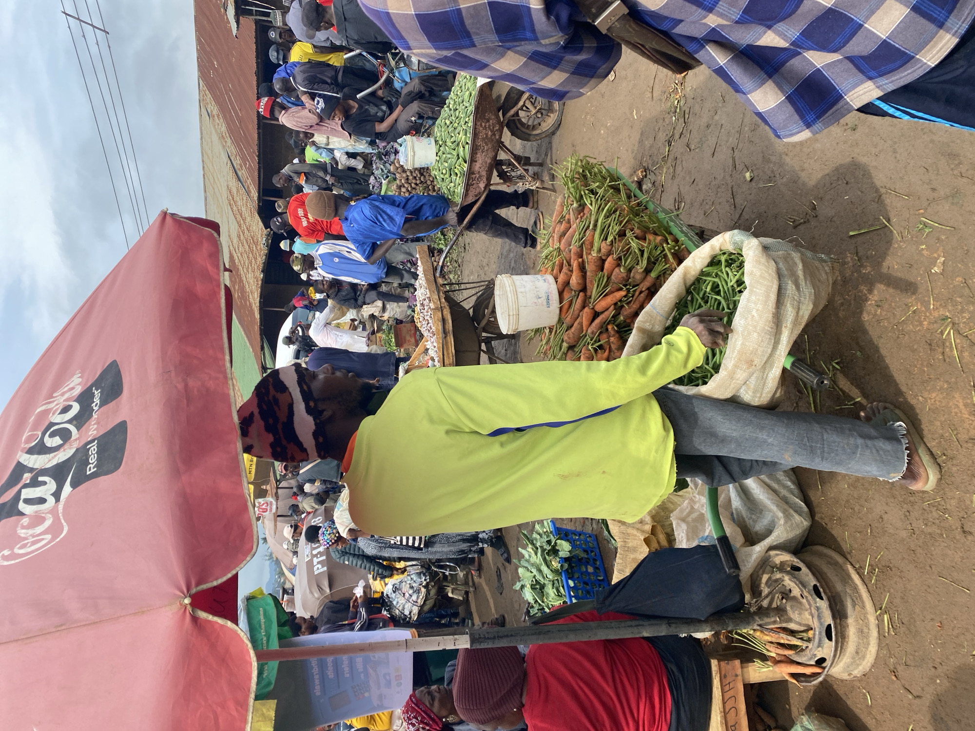 This is a photograph taken at the Katon Rikkos market in Nigeria. The scene depicts a bustling outdoor market with numerous people and stalls. In the foreground, a man wearing a bright yellow long-sleeved shirt and blue jeans is visible from the back, holding a large sack of green beans. Next to him, a cart overflowing with carrots is displayed. Behind him, a large red Coca-Cola umbrella shades a section of the market. Various individuals are seen browsing, selling, and moving about the market. Stalls are set up with produce and other goods, and the ground appears to be dirt. The overall atmosphere is that of a typical Nigerian open-air market, vibrant and full of activity.