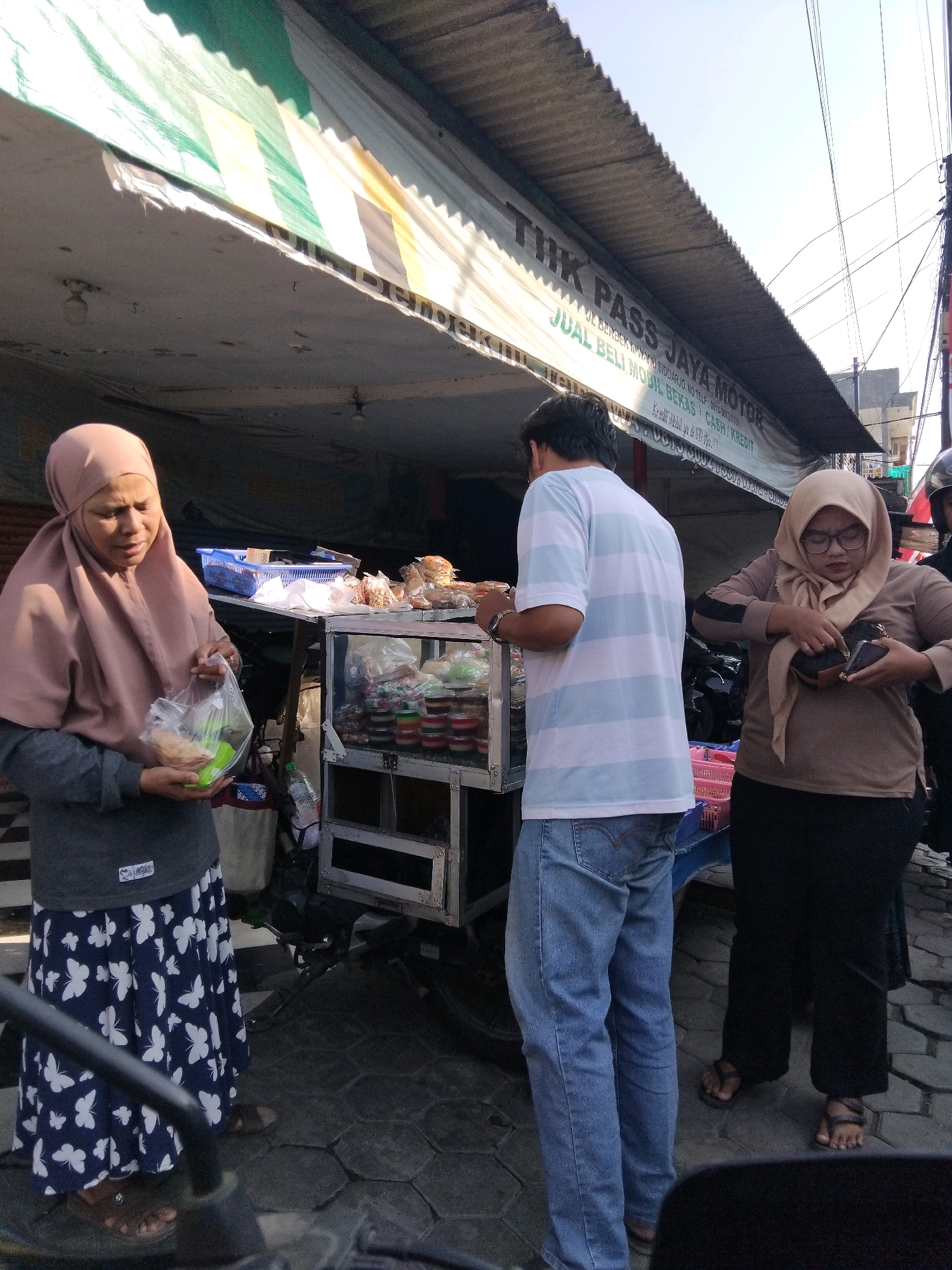 In Wadungasri, Indonesia, a street vendor stands behind a cart displaying various food items in containers and bags. A woman wearing a light brown hijab and a dark grey jacket with a white butterfly pattern on her blue skirt, is holding a clear plastic bag containing food. To her right, a man in a light blue and white striped t-shirt and blue jeans is facing the cart. Another woman, also wearing a hijab, this one a darker brown, and a brown long-sleeved shirt with black pants, is on the far right, looking at her phone. The vendor's cart is positioned under a corrugated metal awning with a striped tarp overhead. The ground is paved with interlocking hexagonal stones.