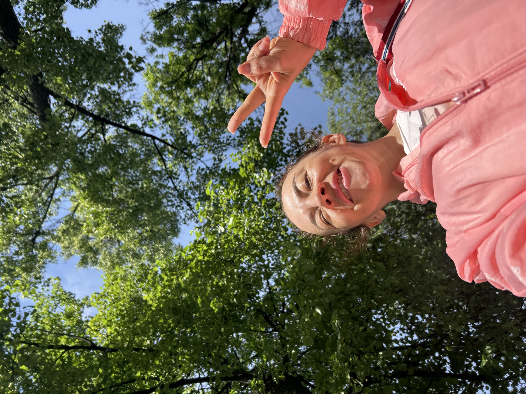 The image is a low-angle shot of a woman wearing a pink jacket making a peace sign with her right hand. Her face is close to the camera, and she is smiling. Behind her is a view of green trees and blue sky. The location is Chaikovsky, Russia.