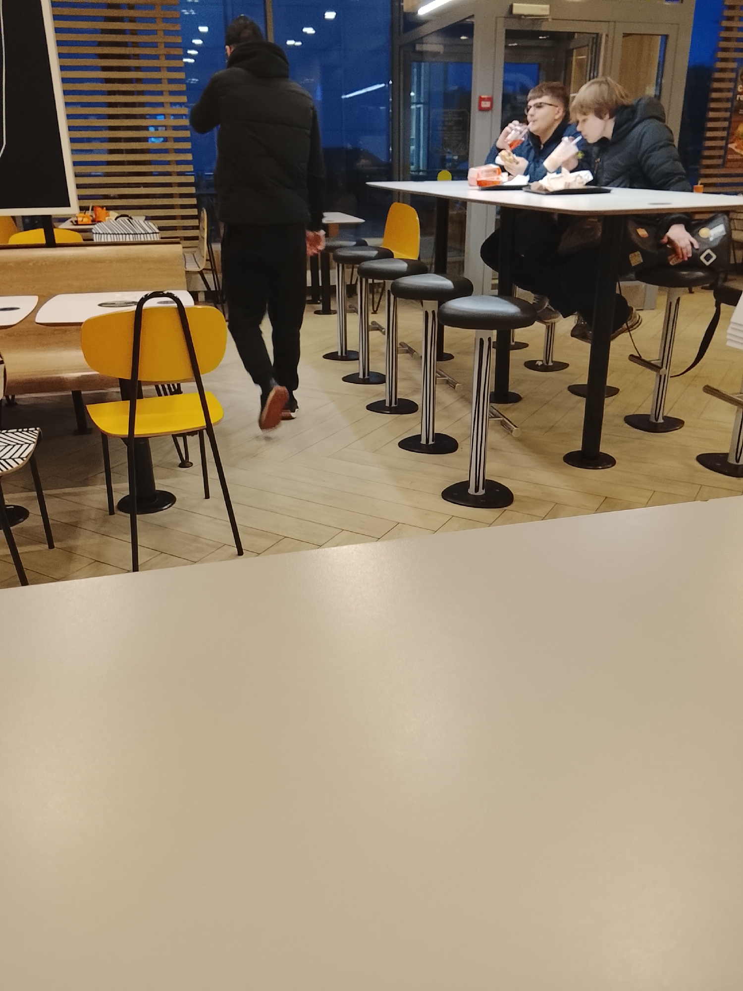The image captures a scene inside a McDonald's restaurant in Obninsk, Russia. Two people, one with glasses, are seated at a white table and appear to be eating. A man in a dark jacket and pants is walking away from the tables, moving from left to right in the frame. The restaurant features modern decor with yellow chairs and bar stools around the tables. Natural light filters through the exterior windows, suggesting it could be dusk or early evening. The foreground is dominated by a section of a beige table.