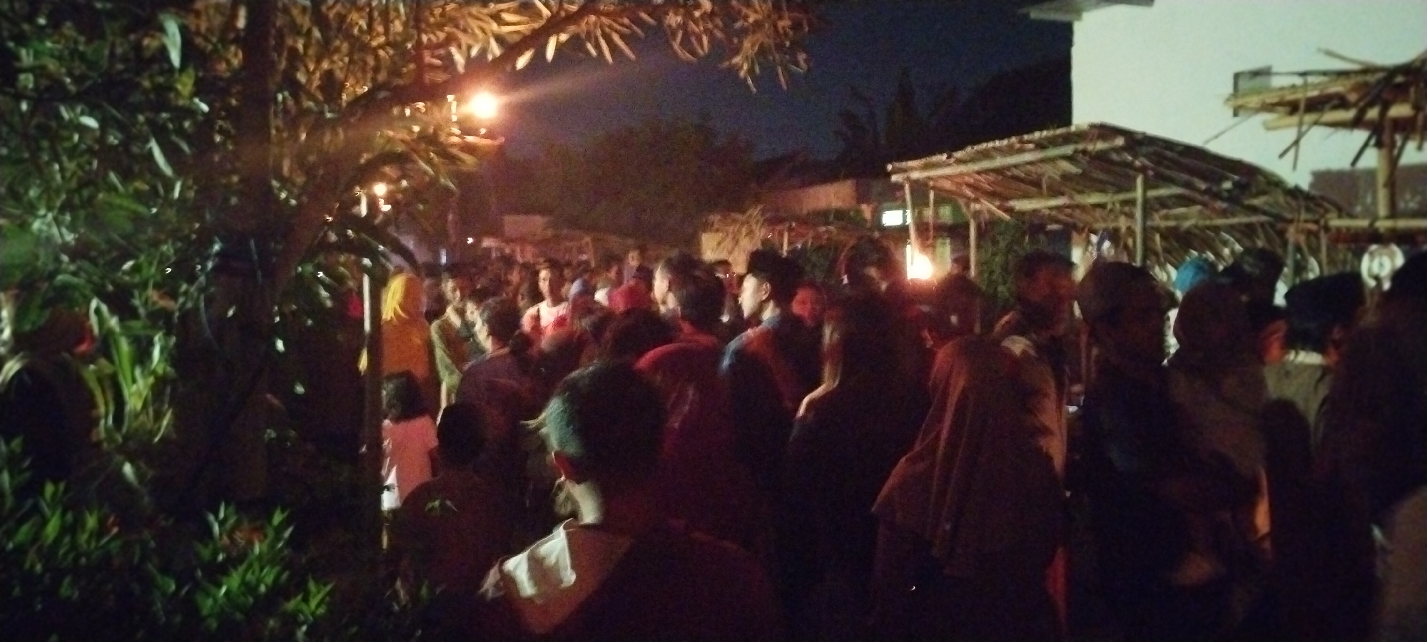 The image captures a dimly lit outdoor gathering at night in Keting, Indonesia. A dense crowd of individuals is visible, many appearing to be of Asian descent. The environment suggests a casual event, possibly a street market or a community gathering, with stalls or temporary structures constructed from wood and thatch in the background. Trees with visible foliage are present on the left side of the frame, and a streetlamp casts a warm glow, illuminating parts of the scene. The overall atmosphere appears active and populated, with people standing and interacting in close proximity. The low lighting conditions contribute to a somewhat grainy and indistinct quality of the image, making it difficult to discern specific facial features or finer details of clothing.