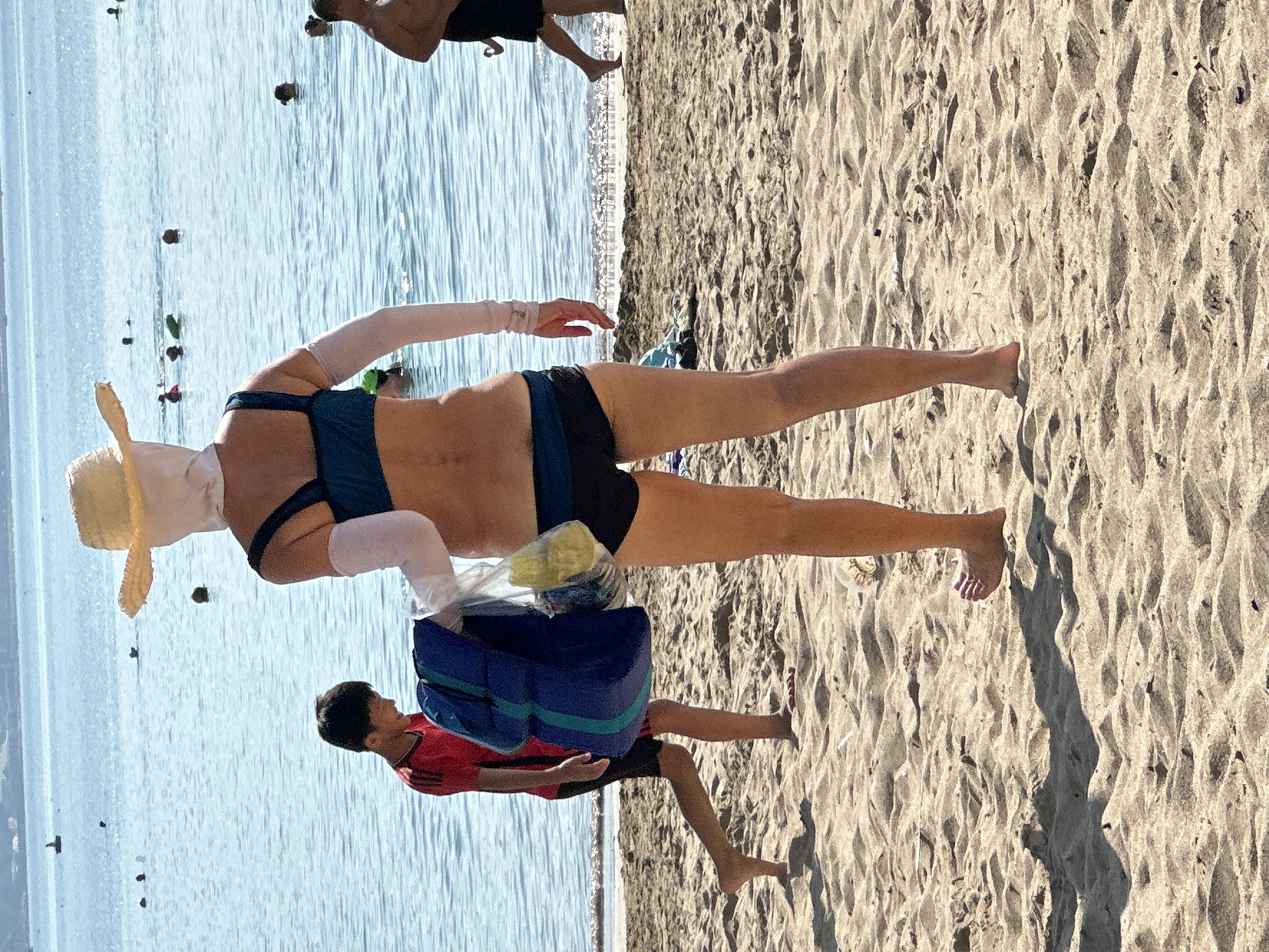The image shows a beach scene, possibly in Bac Nha Trang Ward, Vietnam, with the ocean on the left and sand on the right. An adult wearing swimwear, arm coverings, and a straw hat is walking on the beach. A child, wearing a red shirt and dark shorts, is carrying a blue and green bag. Other people are visible in the water. The beach is sandy with ripples and footprints. The sky is bright.