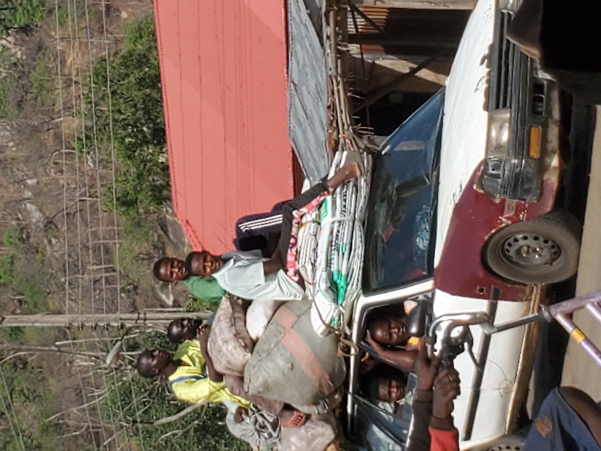 The image depicts a car overloaded with people and goods in Mararaba, Nigeria. Several individuals are crammed inside the vehicle, while others are sitting atop bags and bundled materials loaded on the car's roof. The car itself is a white and maroon sedan. The background shows a hillside, a red-roofed building, and power lines, suggesting an urban or semi-urban setting.