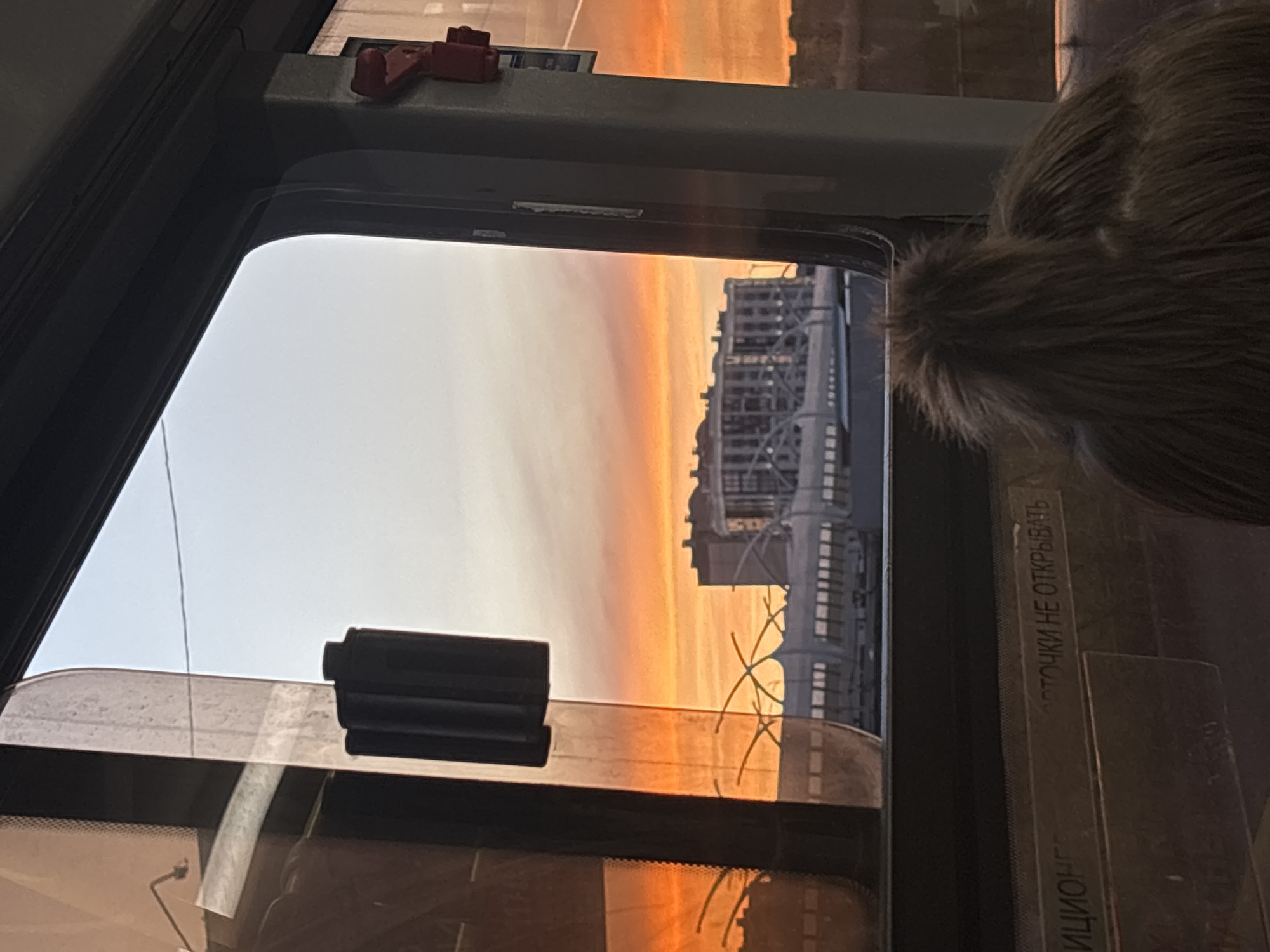 An exterior shot is captured through a window, showcasing a sunset with vibrant orange and pink hues in the sky. In the middle ground, a large, multi-story building with many windows is visible, along with an arched bridge structure. The foreground is occupied by the interior of a vehicle, with a portion of a person's head and hair visible on the right side, and a close-up view of a window frame and a sticker with Russian text that reads "ЗАСТОЧКИ НЕ ОТКРЫВАТЬ" (which translates to "DO NOT OPEN THE WINDOWS"). The reflection on the left side of the window shows the exterior environment.