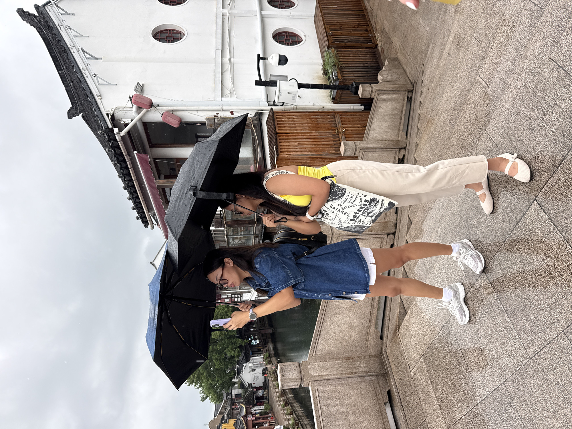 Two women stand on a stone bridge in Qingpu District, China, holding umbrellas to shield themselves from the rain. The woman on the left wears a denim jacket, white shorts, and sneakers, and holds up a smartphone to take a picture. The woman on the right wears a yellow tank top, beige pants, and white flats, and carries a patterned tote bag. Behind them, traditional Chinese architecture lines a canal, with trees and a cloudy sky completing the scene.