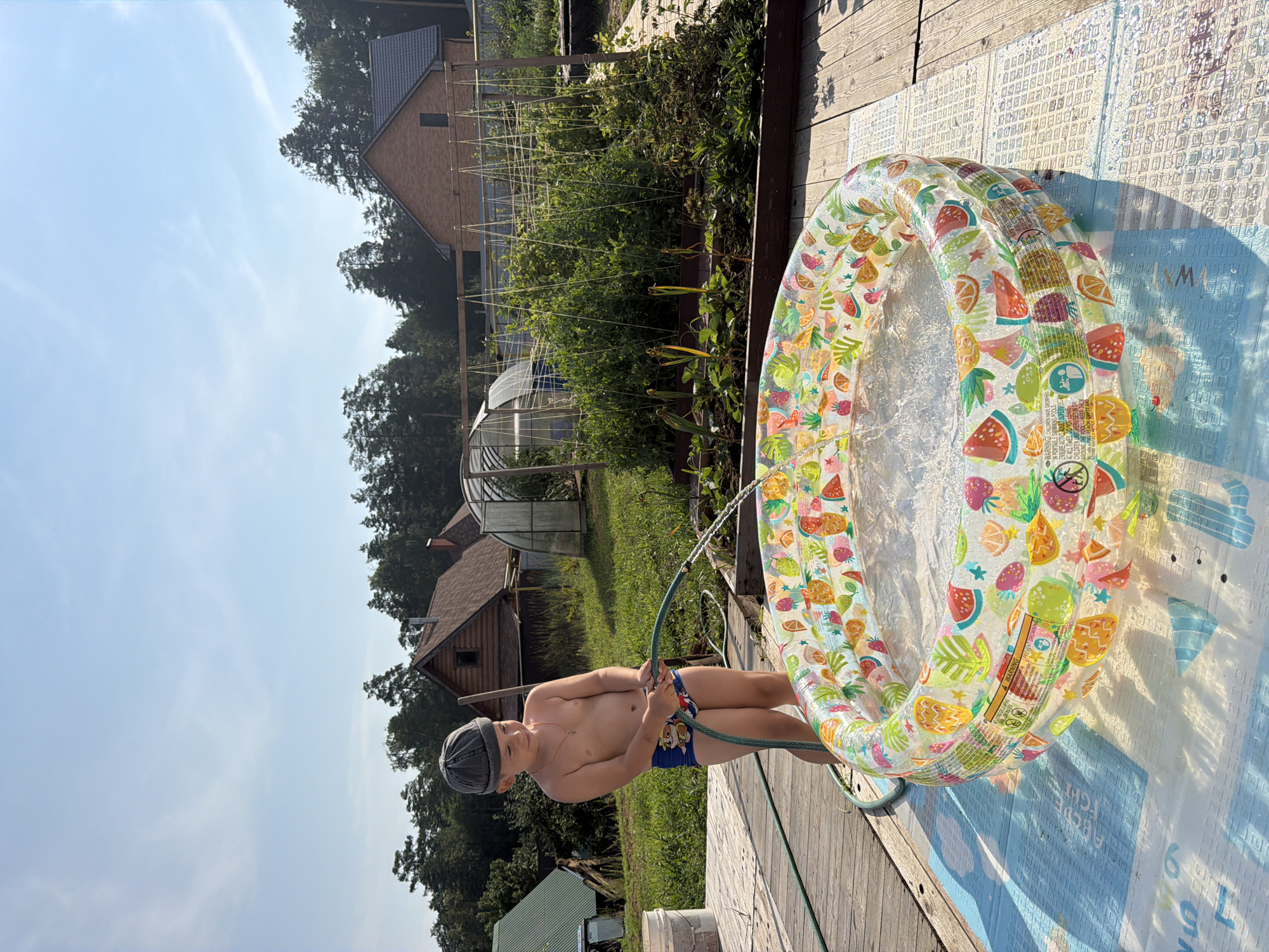 A young boy wearing a grey cap and blue swim trunks is filling a colorful inflatable pool with water from a green hose. The pool has a fruit pattern. He stands on a wooden deck next to a blue mat with alphabet letters printed on it. Behind him, there is a garden with rows of vegetables and a greenhouse. Several wooden houses are visible in the background, surrounded by trees. The sky is clear and blue. The scene appears to be a backyard in a rural or suburban area, possibly in Chaikovsky, Russia, given the architecture and vegetation.