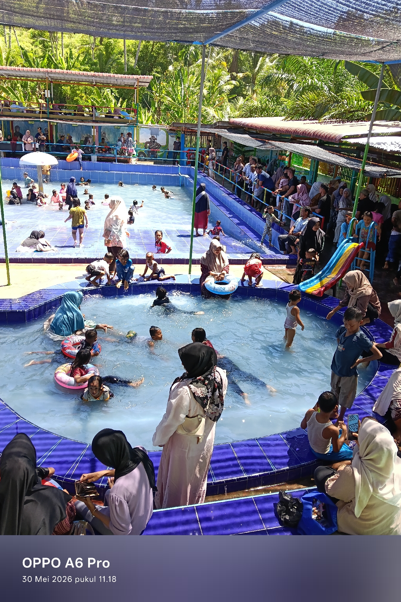 The image shows a crowded swimming pool complex in Palohseulimeng, Indonesia. There are two connected pools, one larger and rectangular, and the other smaller and irregularly shaped. Both pools are filled with people of various ages, including children and adults, some swimming and others wading. Some individuals are wearing swimsuits, while others are wearing clothing and headscarves. Several people are using inflatable tubes. The area surrounding the pools is tiled, with many spectators observing from the edges. A slide is positioned near the edge of the pool. The background includes lush greenery, trees, and various buildings or structures, indicating an outdoor recreational setting. Overhead netting provides some shade. The bottom of the image shows camera data indicating it was taken with an OPPO A6 Pro.