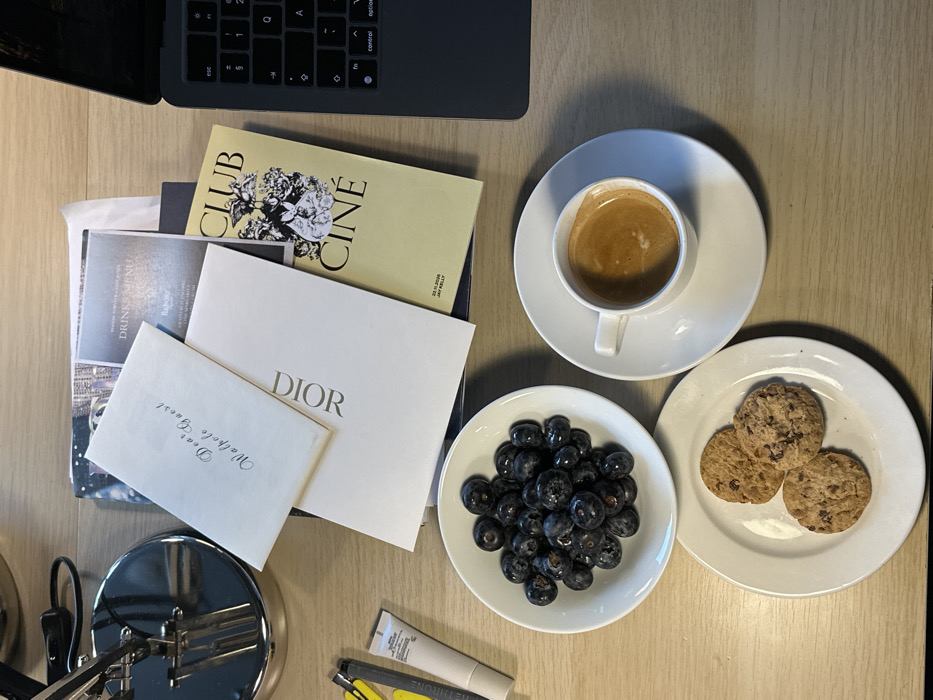 The image displays an overhead view of a light wooden desk surface. In the foreground, a white bowl holds numerous blueberries, and a white plate contains three chocolate chip cookies. To the right of the blueberries, a white espresso cup with visible crema sits on a matching white saucer. In the background, a stack of papers and booklets is visible. The uppermost item is an off-white card printed with "Dear Walpole Guest." Directly beneath it lies a white card embossed with "DIOR" in gold lettering. Partially visible below these are a "DRINKS MENU" and a light yellow booklet titled "CLUB CINÉ" with floral graphics and the inscription "22.11.2020 JAY KELLY." To the far upper right, a dark grey laptop keyboard is partially visible. On the upper left, a metallic desk lamp is partially seen, alongside a yellow utility tool and a small white tube of cream. The scene indicates a luxurious setting, likely a high-end hotel room or private club in London, United Kingdom, accommodating a guest associated with the Walpole Group, the official sector body for British luxury. No individuals are present in the image.