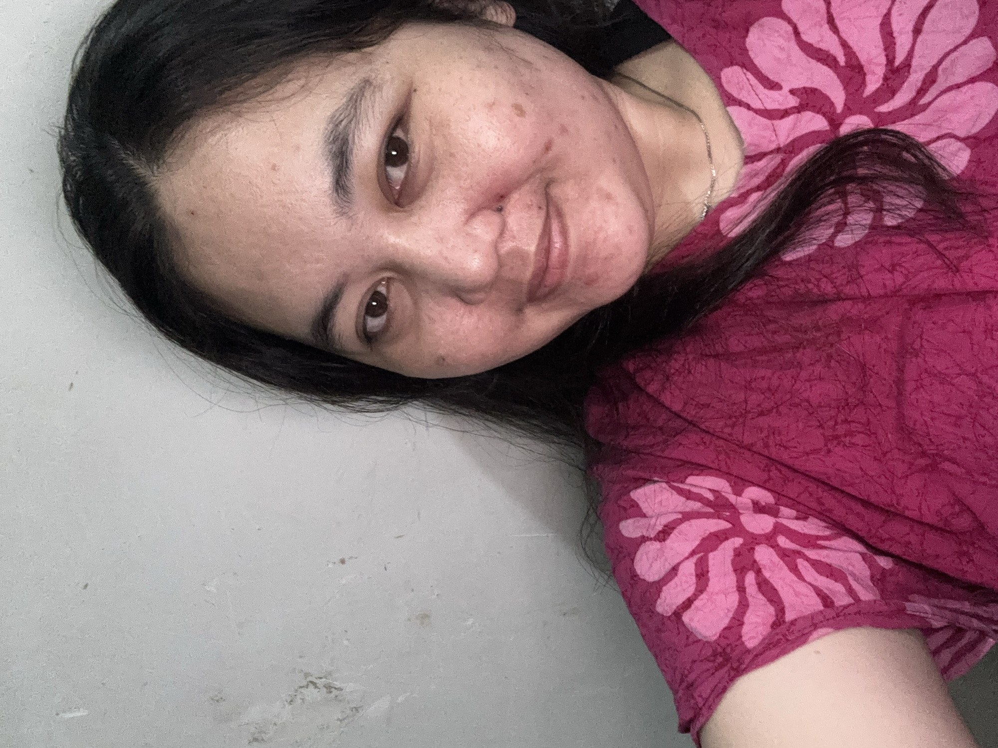 A close-up, waist-up selfie shows a young woman with dark hair and brown eyes posing in front of a grey, textured wall. She is wearing a vibrant pink t-shirt with a large, lighter pink floral pattern. The woman has a light tan complexion with some visible blemishes and freckles. She is smiling gently, with her head tilted slightly to her left. A thin silver necklace is visible around her neck. The background wall appears to be an interior surface, possibly a painted concrete or plaster wall with some signs of wear and tear. The lighting suggests an indoor setting, possibly a bedroom or living space. No specific location in Unknown, Indonesia can be determined from the image.