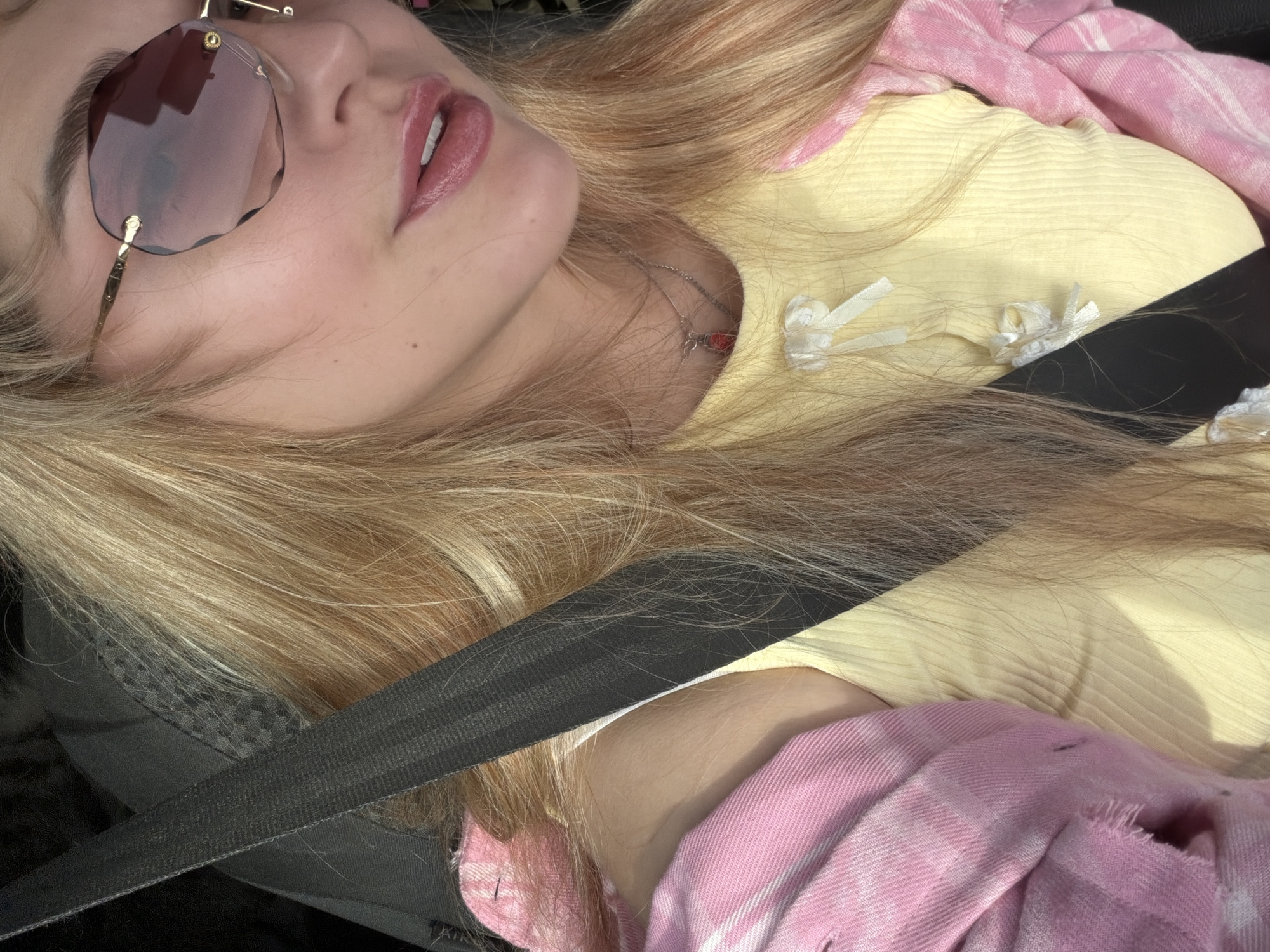 A close-up, high-angle shot captures a person, likely female, with long, light blonde hair, reclining in what appears to be a car during the daytime. She is wearing large, rimless sunglasses with reddish-tinted, irregularly shaped lenses and delicate gold-colored accents. Her lips are slightly parted, adorned with a glossy, pink-toned lip product.
The subject is dressed in a light yellow ribbed top, which features small white bow-like details on the chest. A pink and white plaid shirt or jacket is worn over or draped around her shoulders and upper body. A thin silver-colored necklace with a small red pendant is visible around her neck. A black seatbelt strap diagonally crosses the lower left portion of the image, indicating she is a passenger in a vehicle. Portions of a black car seat or interior are also visible. The natural, bright lighting suggests the image was taken during the day. The person appears relaxed, with no specific activity occurring beyond resting in the car. There is no visible text in the image.