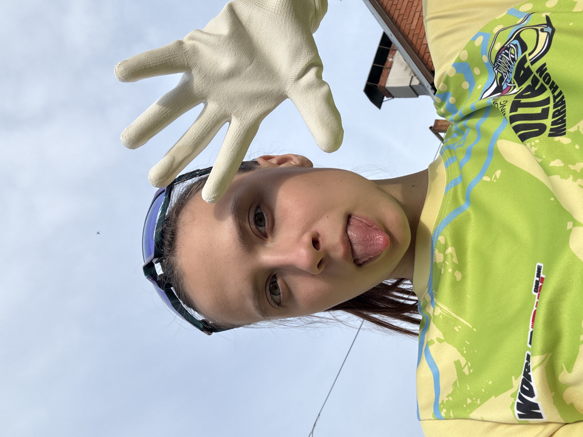 A young woman is pictured in a low-angle shot, sticking out her tongue. She is wearing sunglasses and a yellow and green shirt that reads "ULTRA MARATHON" and "WORLD ONLINE". Above her head is a hand wearing a white glove. The background shows a blue sky and part of a building with a brown roof. The image was taken outdoors in Прикамский, Russia.