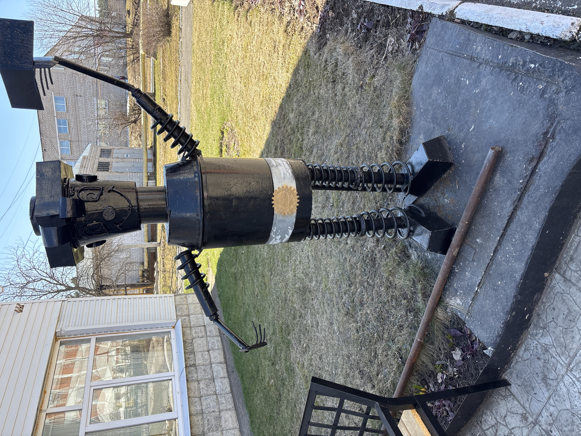 The image depicts a black metal sculpture resembling a robot, standing on a concrete base outdoors. The sculpture is made of various metal components, including a cylindrical body with a silver band, spring-like legs and arms, and a head made of rectangular shapes. The sculpture holds a rectangular object over its head and has a small rake attached to one of its arms. The setting appears to be a park or outdoor area in the city of Chaikovsky, Russia, based on the style of architecture in the background, which includes a white building with a large window and a brick building. The ground is covered with grass and some patches of dry vegetation. A metal bench frame is partially visible in the foreground. The sculpture is set on a concrete base, next to which lies a metal rod.