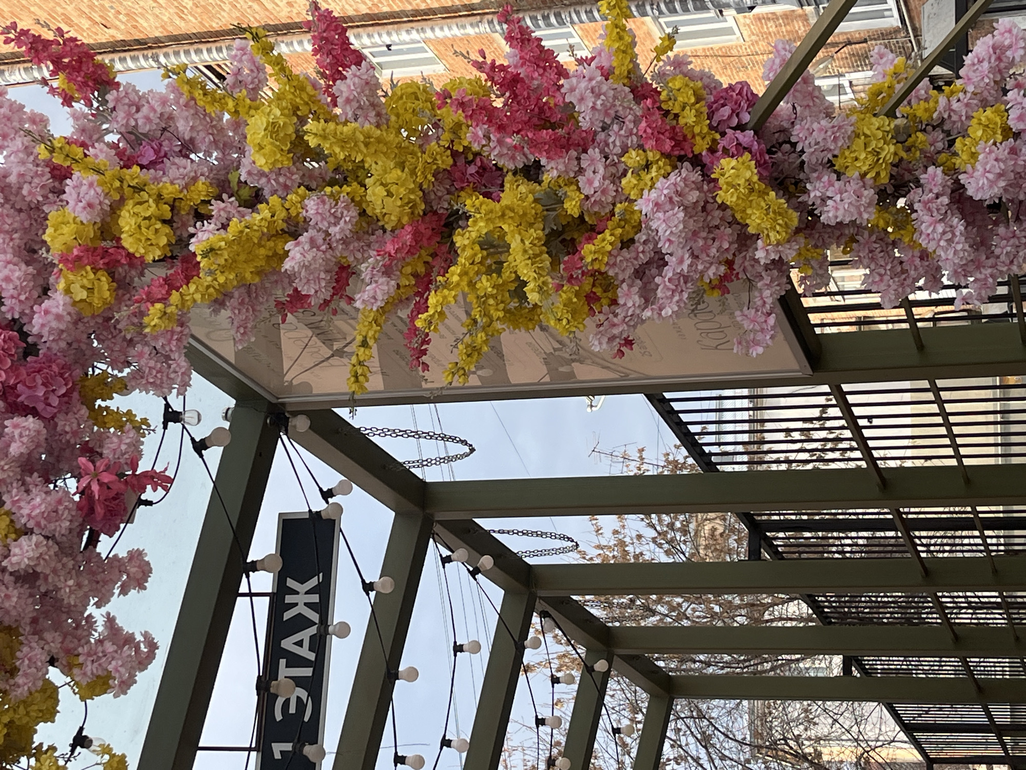 The image depicts a street scene in Izhevsk, Russia. An ornamental archway is decorated with abundant artificial pink, yellow, and red flowers, positioned over what appears to be an entrance or pathway. The archway is constructed of a green metal framework, adorned with strings of lights. A sign with Cyrillic text ("1 ЭТАЖ") hangs from the structure. The upward angle provides a view of the sky and building facades. The architectural details indicate an urban environment.