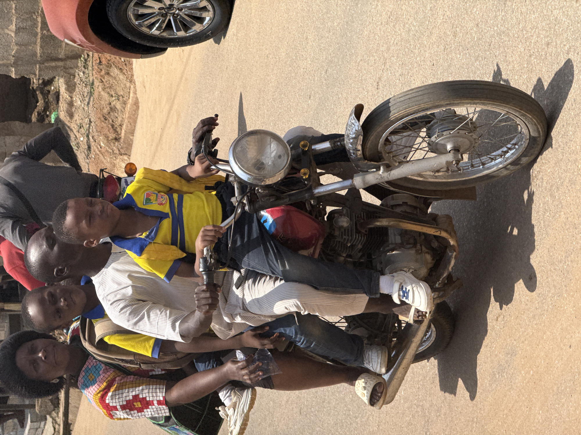 This media file captures a group of people on a motorcycle in Abuja, Nigeria. The scene appears to be outdoors on a paved surface, likely a street or road, with a dirt area visible to the left, suggesting an urban or semi-urban environment. The lighting indicates it is daytime, with strong shadows cast by the subjects and the motorcycle, hinting at a sunny day.
The central focus is a motorcycle with at least four individuals on it. In the front, a young person in a yellow and blue shirt appears to be controlling the motorcycle. Behind them, two more young people are seated, and a woman is positioned at the very back. The people are dressed in casual clothing. One of the young passengers wears a white and striped shirt, and another wears jeans. The woman at the back is wearing a colorful, patterned garment.
The motorcycle is an older model, visible with its engine, handlebars, headlight, and exhaust pipe. The overall impression is a common mode of transportation in the depicted location, possibly carrying individuals to a destination or engaged in travel within the city. The expressions on the faces are not clearly discernible to infer specific emotions, but the arrangement suggests a functional use of the motorcycle for group transit. No visible text is present in the image.