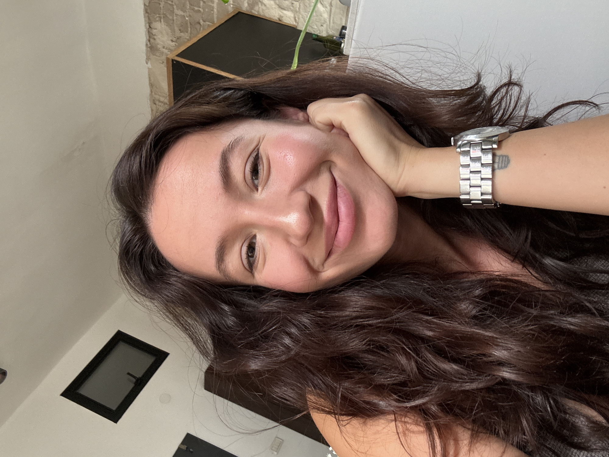 A woman with long, dark, wavy hair smiles at the camera, resting her chin on her hand. She wears a silver watch on her left wrist. The background shows a white wall with a black framed window at an angle, and some exposed brickwork. The scene suggests an indoor setting, possibly an apartment or studio, with natural light contributing to the subject's glow. There is no indication of the city of Tel-Aviv, Israel.