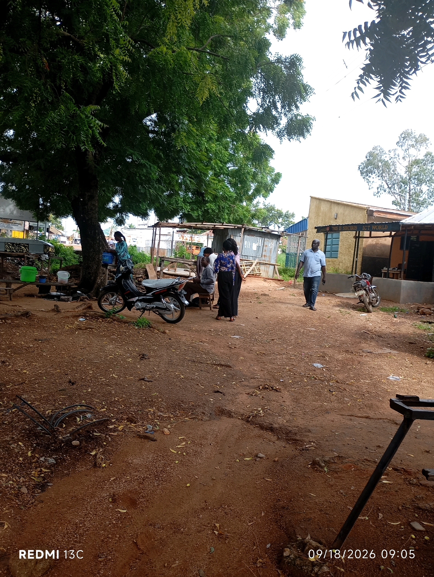 The image depicts an outdoor scene, likely a market or roadside stall area, in Shendam, Nigeria. A large tree with lush green foliage dominates the left side of the frame. In the foreground, reddish-brown dirt ground is visible, scattered with fallen leaves and small stones.
Several individuals are present. A woman in a blue and green patterned outfit stands next to a parked motorcycle under the tree, holding a blue bucket. A second woman, wearing a dark, patterned top and a long black skirt, stands to her left, with her back to the camera. Further into the scene, a man in a light blue shirt and dark pants walks towards the right, his gaze directed forward. Two other individuals are seated on a bench or low structure near the background.
Objects in the scene include a black motorcycle parked under the tree, and another red motorcycle parked near a building in the background. Various structures, appearing to be makeshift stalls or small buildings, are visible behind the people. One building on the right has a sign that reads "THIS IS SIRCHRIS ICT WORLD". In the middle ground, there are wooden benches and tables, some with green buckets and other containers. In the far left, some metal frames are stacked.
The overall impression is a busy, functional outdoor space, likely a commercial area within the city. The sky is bright and overcast, suggesting daytime.