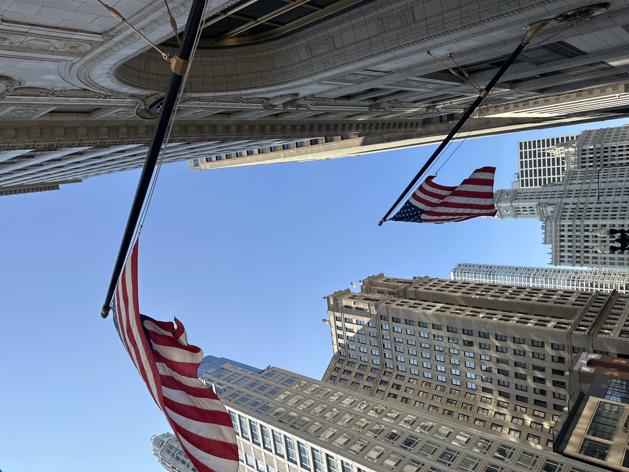 In this low-angle shot of Chicago's Loop, two American flags are prominently displayed against a clear blue sky. The flag on the left is positioned at the top of the frame, its red and white stripes billowing. Below it, a series of tall buildings, characterized by their grid-like window patterns and varying architectural styles, dominate the cityscape. One building, in particular, features a lighter, more ornate facade with arched windows and decorative detailing.
To the right, another American flag extends from the side of a grand, light-colored stone building. This building's facade is highly textured with relief carvings and features large arched windows. Further in the distance, the iconic clock tower of the Wrigley Building is visible, along with other skyscrapers that define the Chicago skyline. The overall scene captures a classic urban environment, highlighting the juxtaposition of architectural grandeur and national symbolism under a bright sky.