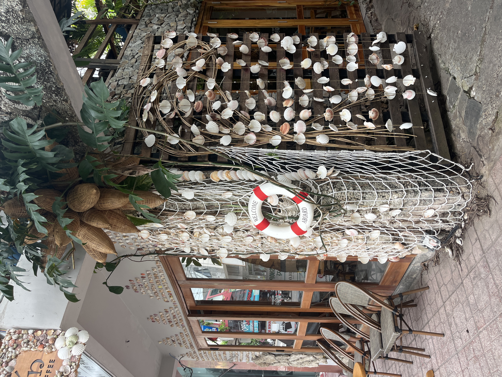 A medium shot captures the exterior of a coffee shop in Nha Trang, Vietnam, decorated with a nautical theme. The building's facade is adorned with numerous seashells, and a fishing net hangs from a tree, draped with more shells. A life preserver with "Welcome Aboard" written on it is attached to the net. In front of the shop, several empty wicker chairs with brown frames are arranged on a tiled patio. Wooden doors with glass panes lead into the establishment. The scene is devoid of people.