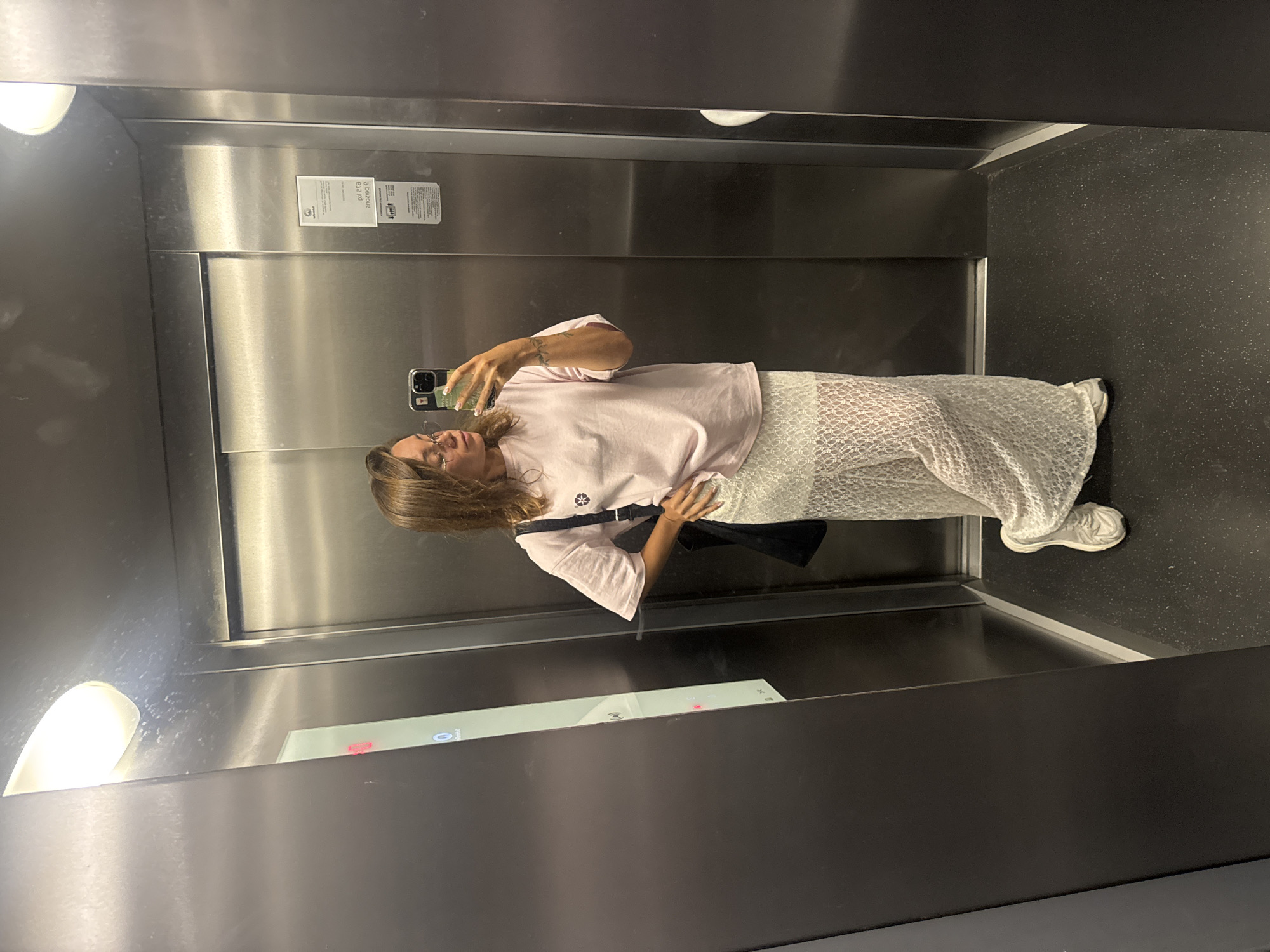 The image captures a woman taking a selfie in a modern elevator. She is reflected in the polished stainless steel wall, holding a smartphone with a light-colored, patterned case up to capture her image. The woman has light brown, shoulder-length hair and is wearing glasses. She is dressed in a light pink short-sleeved top, a long white skirt with a delicate lace or textured pattern, and white sneakers. A black bag is visible, slung across her body.
The elevator interior features clean, reflective stainless steel walls, a dark, possibly speckled, floor, and integrated lighting fixtures on the ceiling and side panels. A white notice is affixed to the wall behind her reflection, displaying mirrored text, with fragments like "Bezdouz" and "0.12 m3" discernible among other unreadable characters, likely elevator specifications or safety information. A digital screen or control panel is partially visible below her reflection, showing some unidentifiable icons or indicators. The setting suggests a contemporary building in Budva, Montenegro. The mood is casual, depicting a common modern activity.
