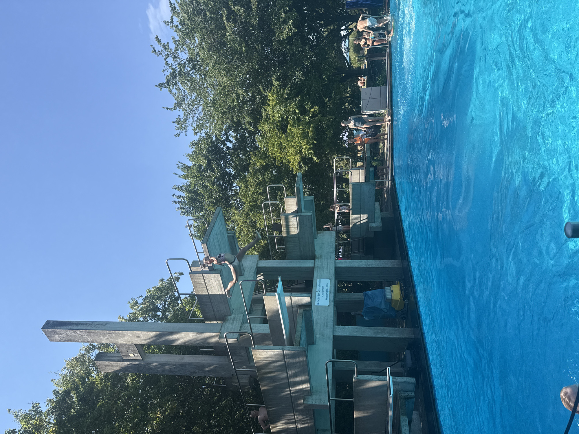 In Rheinbach, Germany, a high diving platform at an outdoor swimming pool is the central focus. The concrete structure features multiple levels and diving boards. A young person in a swimsuit is captured mid-air, having just jumped from one of the upper boards, their body arched in a diving pose. Below, the bright blue water of the pool is visible, with ripples indicating recent activity. Several other individuals are present around the pool, some standing on the edge and others in the water. Trees form a lush green backdrop against a clear blue sky.
