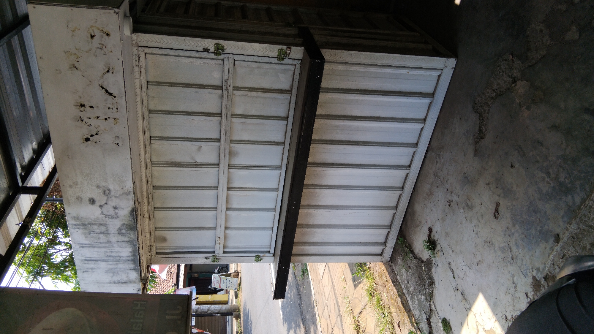 The image shows a weathered, white corrugated metal kiosk with a dark, shelf-like counter. The kiosk has vertical panels and is situated on a concrete surface in what appears to be an outdoor urban environment. Some green vegetation is visible growing through cracks in the concrete. In the background, a street can be seen with a motorcycle parked and a sign with Indonesian text. The overall impression is one of a modest, potentially abandoned or disused commercial structure, common in many Indonesian cities like Proyonangg.