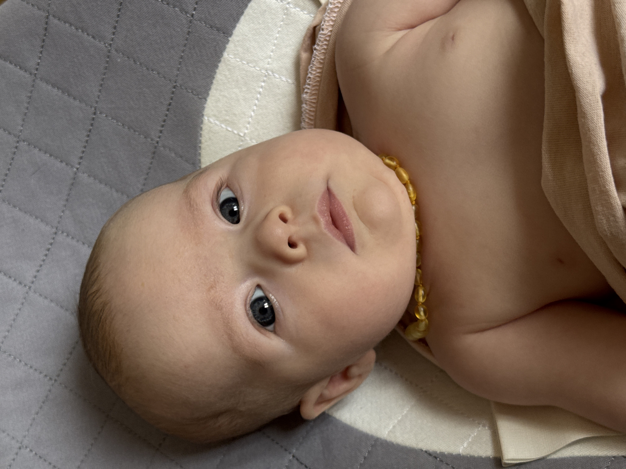 A baby with light skin, dark eyes, and wearing a yellow beaded necklace lies on its back on a quilted grey and white blanket. The baby is partially covered by a beige cloth. The location is not discernible from the image.