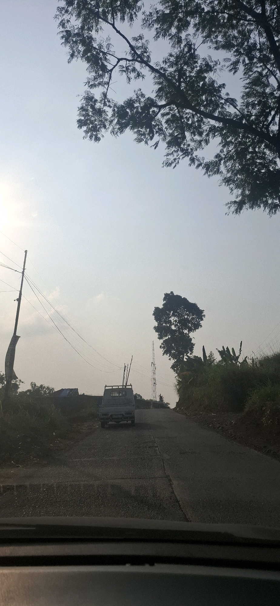The image depicts a rural road scene in Cibinong Hilir, Indonesia, under a bright, overcast sky. In the foreground, the dashboard of a vehicle is visible, suggesting the photo was taken from inside a car. The road itself is paved with visible seams and appears to be in fair condition, with a slight incline. A light-colored pickup truck, loaded with long, thin poles or pipes in its bed, is driving away from the camera up the road.
To the left of the road, a utility pole with several wires stretches upwards, and a large banner or flag hangs from it, partially obscured by the truck. Further back on the left, there are some structures with blue tarps, possibly temporary shelters or storage. The landscape to the left is covered in vegetation and appears somewhat overgrown.
On the right side of the road, a steep embankment rises, covered in dense green foliage, including what appear to be banana plants. Dominating the background is a tall telecommunications tower. A large, solitary tree with a full canopy stands prominently near the tower and on the embankment. Overhead, the branches of a large tree, silhouetted against the sky, frame the upper portion of the image. The overall impression is of a tranquil, somewhat undeveloped area.