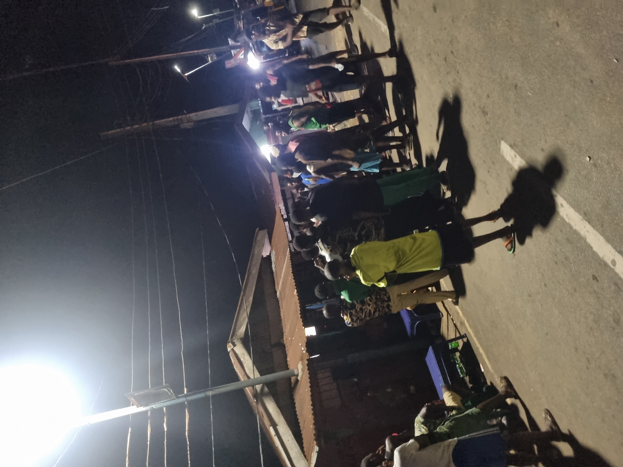 A dimly lit outdoor scene, possibly at night, in the city of Onitsha, Nigeria, shows a group of people gathered together. They appear to be standing and sitting along the side of a road or street. The area is illuminated by overhead streetlights, which cast shadows of the people onto the pavement. The subjects appear to be a mix of adults and children. The environment includes a building with a corrugated metal roof, streetlights, power lines, and what appears to be a paved street. The details about the subjects are limited due to the image quality and lighting.