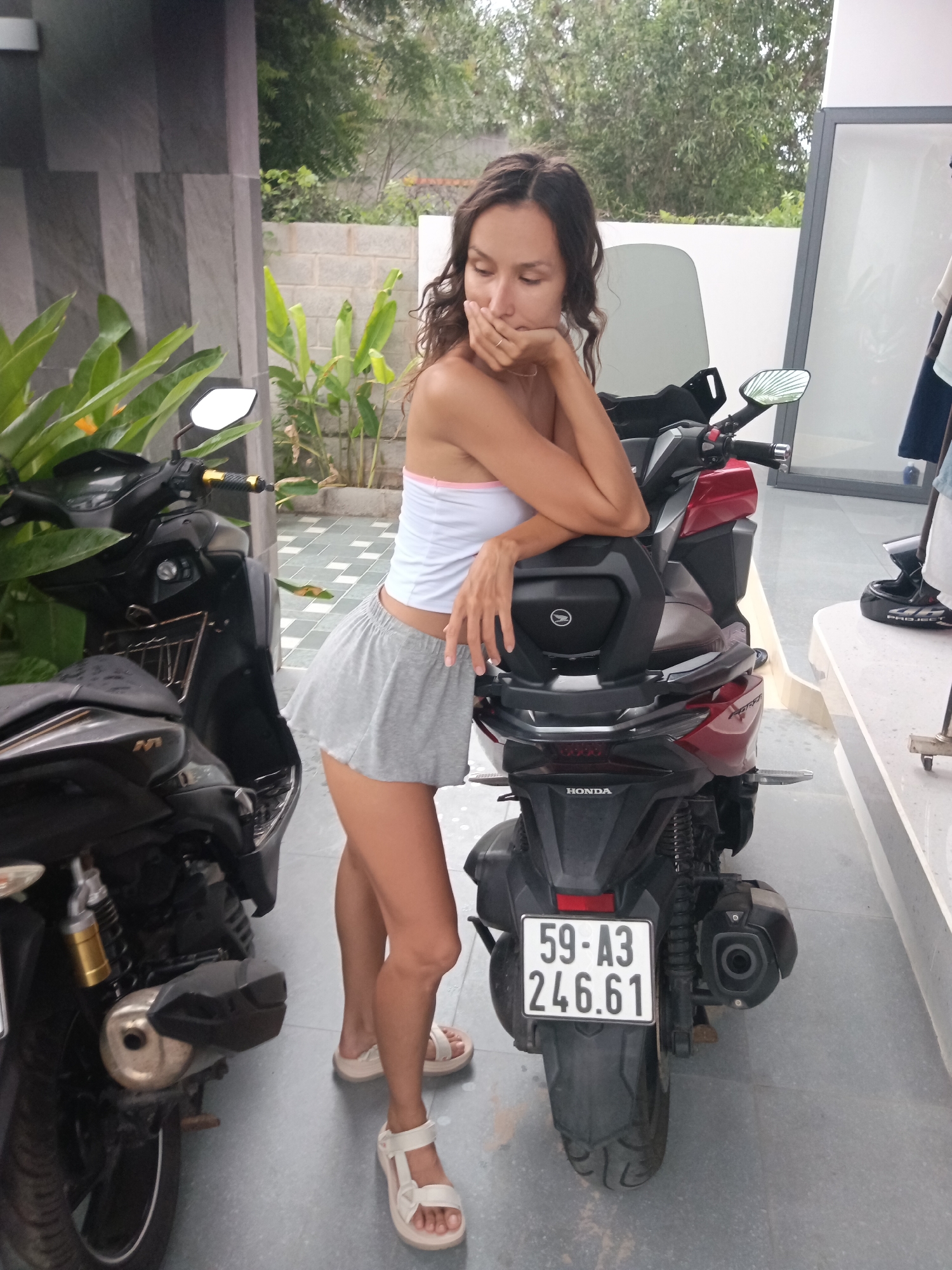 A woman with long brown hair is standing next to two motorcycles. She is wearing a white bandeau top with a pink trim and a grey pleated mini skirt. Her right arm is bent and resting on the seat of the red motorcycle, with her hand covering her mouth. She is wearing white sandals. The motorcycles are parked on a tiled patio. In the background, there is a building with grey striped walls and lush green plants. To the right of the red motorcycle, a white counter with a grey edge is visible. The scene appears to be set in Mũi Né, Vietnam.
