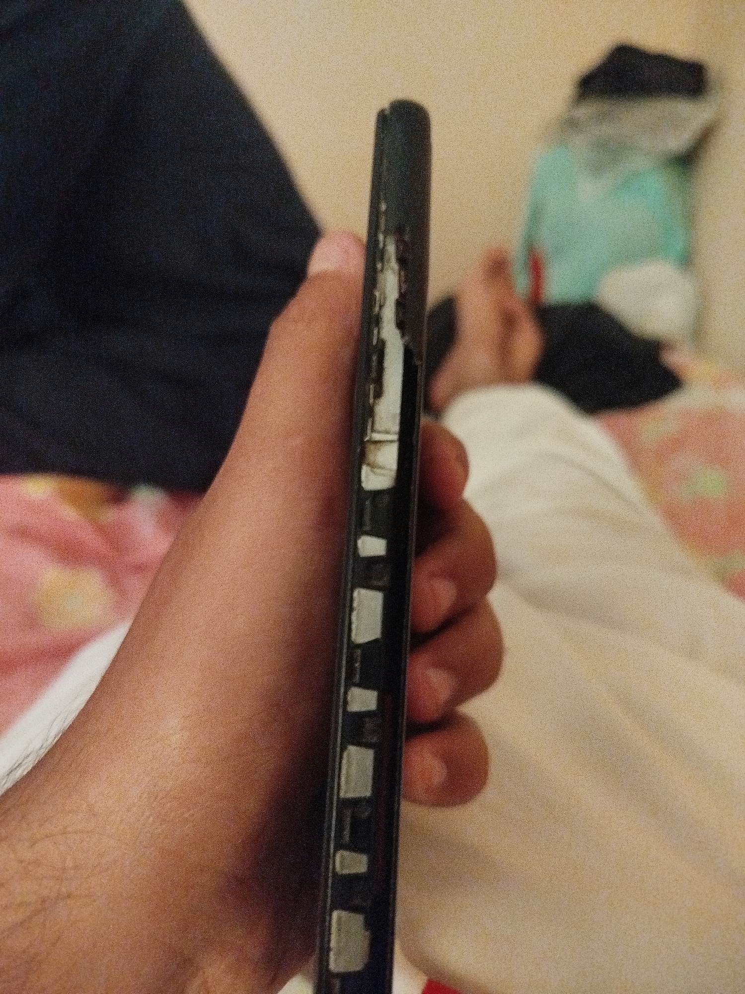 The image shows a close-up of a hand holding a damaged black smartphone. The phone's casing is peeling away, revealing white components underneath. The background is blurred and appears to be a bedroom with a person lying on a bed, covered by blankets. The lighting is dim. There is no indication of the location being in Lassan, Pakistan.