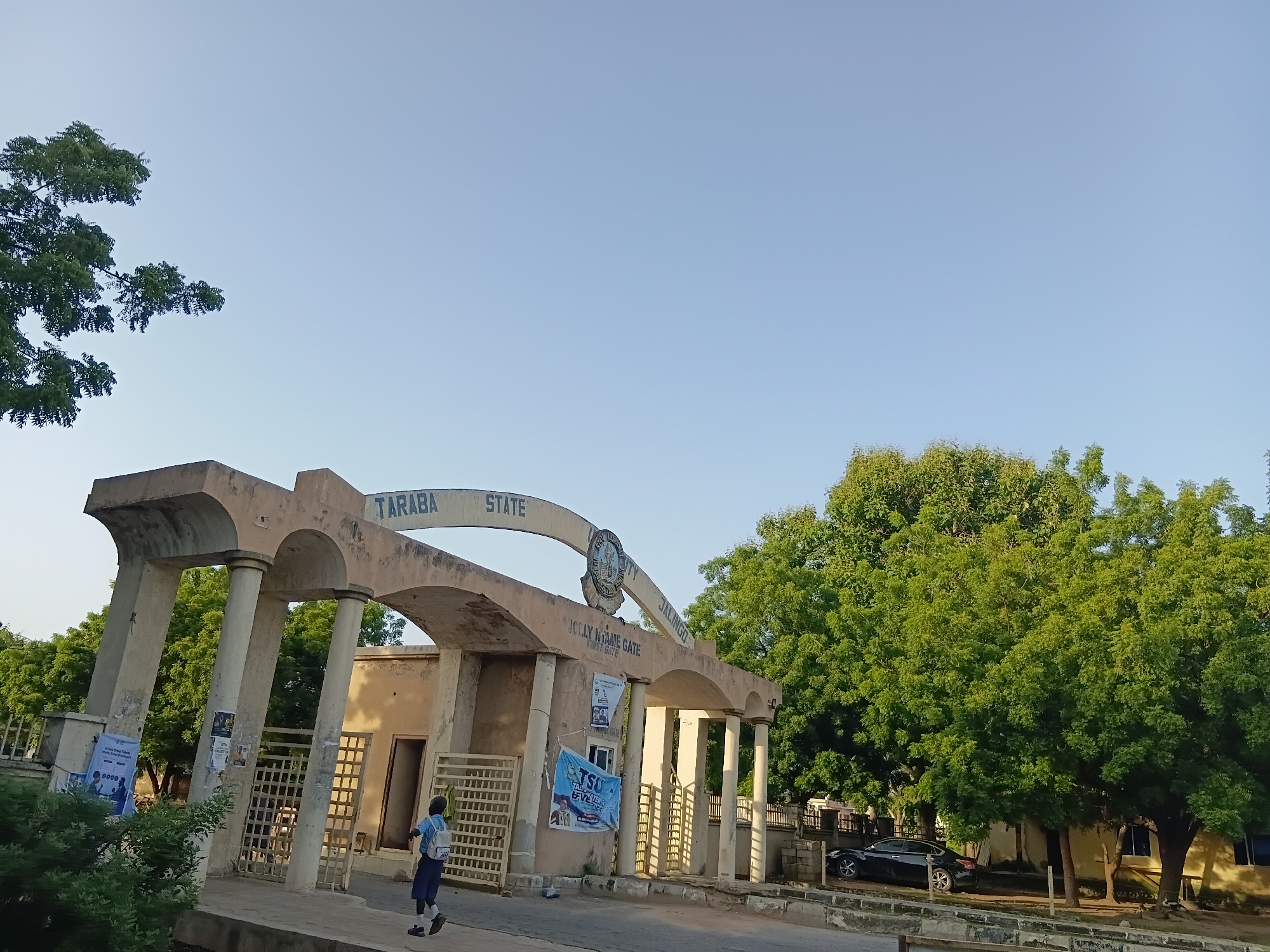 The image shows the entrance gate of Taraba State University in Jalingo, Nigeria. The gate is an archway with several columns. Above the arch, the words "TARABA STATE UNIVERSITY JALINGO" are visible, along with a crest. The gate is made of concrete and appears to be old. There are some posters and banners attached to the gate. A young person in a school uniform is walking through the gate. A black car is parked to the right of the gate, near some trees. The sky is clear and blue. The environment is lush with green trees.