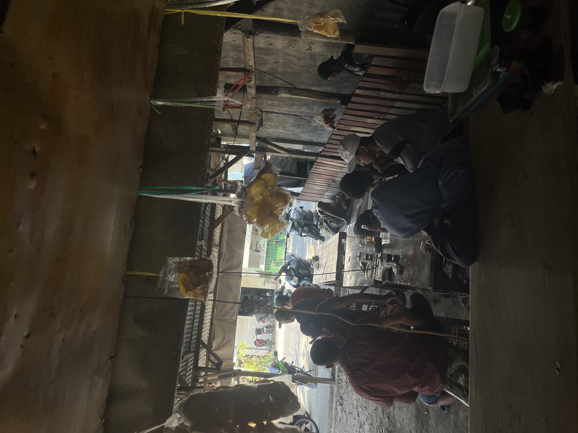 The image depicts an outdoor seating area, possibly a warung kopi, in Dusun Metatu, Indonesia. The scene is partially covered by a corrugated metal roof and a wooden structure. Several individuals are seated at tables, some engaged in conversation, others appearing to rest or observe. Food items, possibly snacks, are displayed in plastic bags hanging from the ceiling. In the background, motorcycles are parked, suggesting a roadside establishment. The overall atmosphere appears relaxed and casual, with evidence of everyday life unfolding. The setting is rustic, with concrete and wooden elements dominating the architecture. The lighting suggests it is daytime, with sunlight filtering through gaps in the roof and the open front of the establishment.