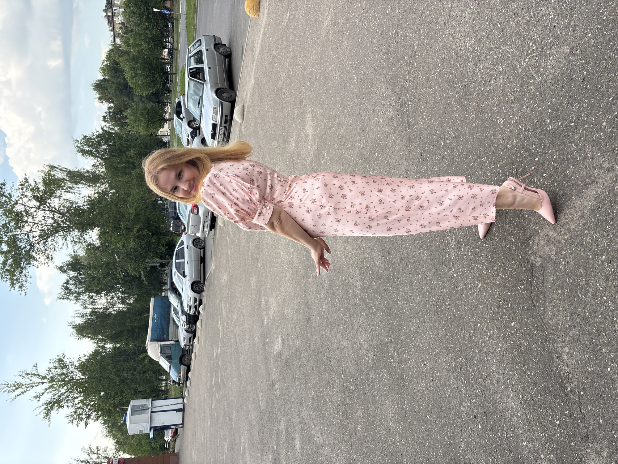 A young woman with blonde hair, wearing a pink floral dress and pink high heels, stands in a parking lot. She is looking back over her shoulder and smiling at the camera. The parking lot is paved with asphalt and is lined with parked cars. In the background, there are trees, a building with a blue and white structure, and a cloudy sky. The image was taken in Yaroslavl, Russia.
