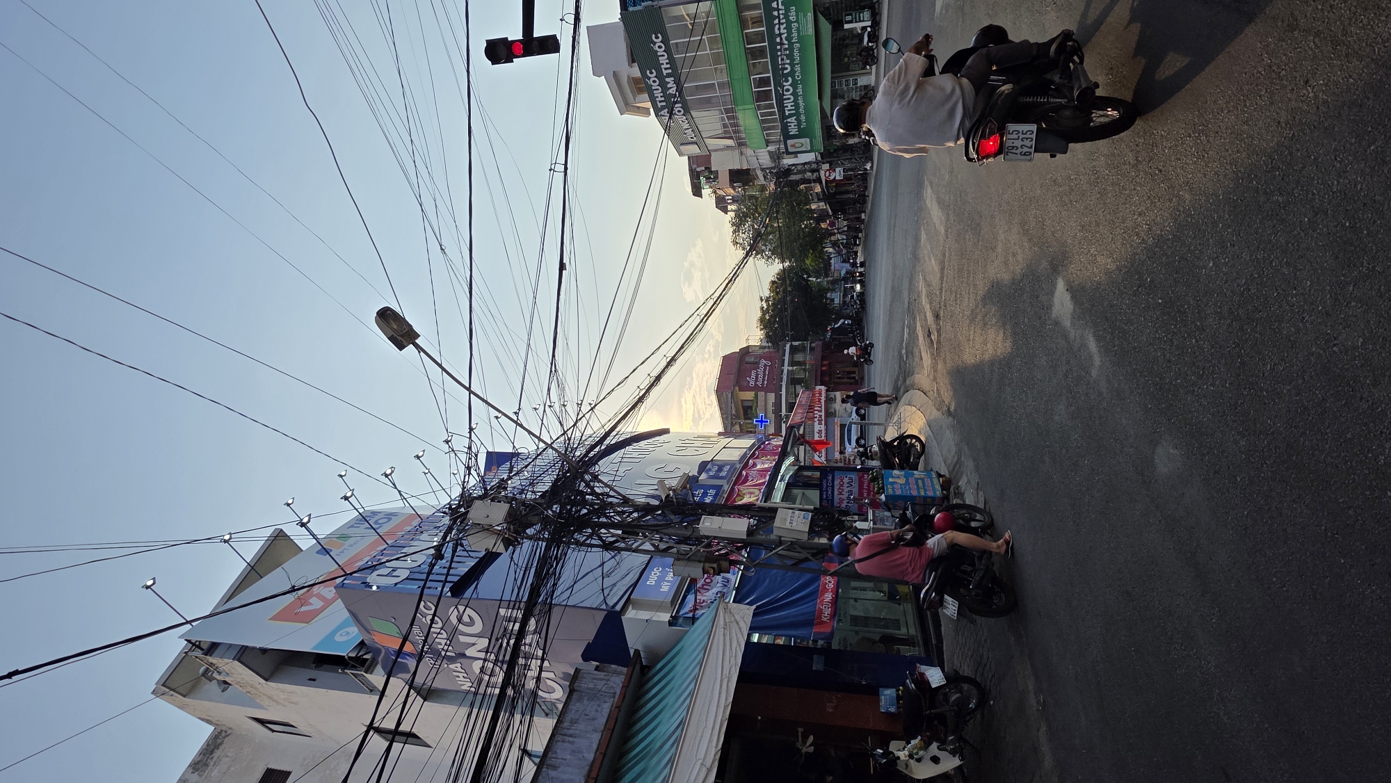 The image depicts a street scene in Nha Trang Ward, Vietnam, characterized by a dense web of overhead electrical wires and a mix of commercial establishments. A red traffic light is visible at the top of the frame. Street-level businesses and signs are visible on both sides of the road, many written in Vietnamese. A motorcycle is driven on the road and other motorcycles are parked along the street. People are present on the road and sidewalk. The sky is pale blue, suggesting it is either early morning or late afternoon.
