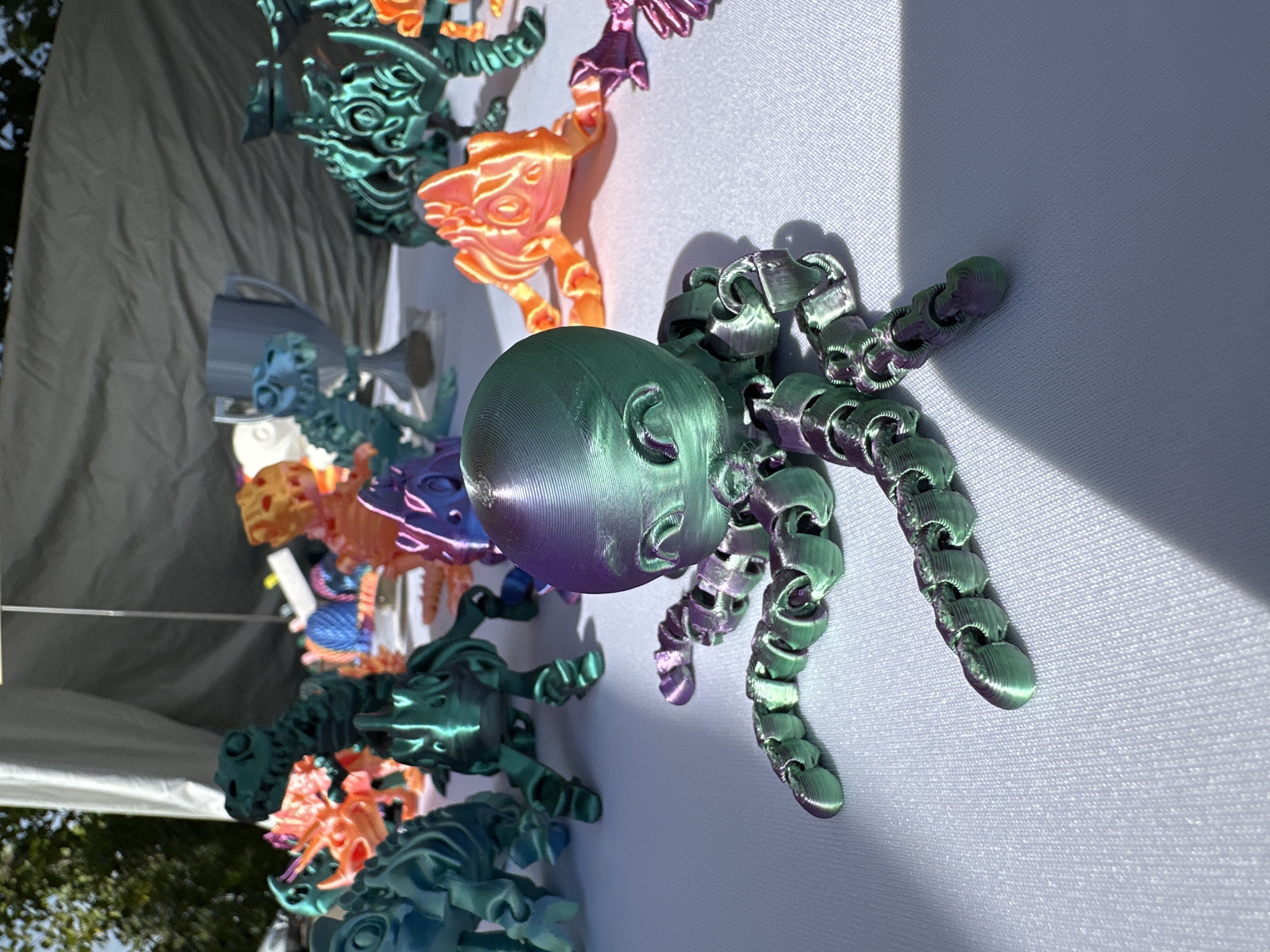 This image shows a table covered with a white tablecloth, displaying several 3D-printed toys, most of which appear to be stylized dinosaurs and an octopus in the foreground. The toys are made from filament that shifts between green and purple. In the background, there are trees, suggesting an outdoor setting, possibly a market or fair. A white tent structure is visible behind the toys. The image does not provide any information about the city of Vilnius, Lithuania.