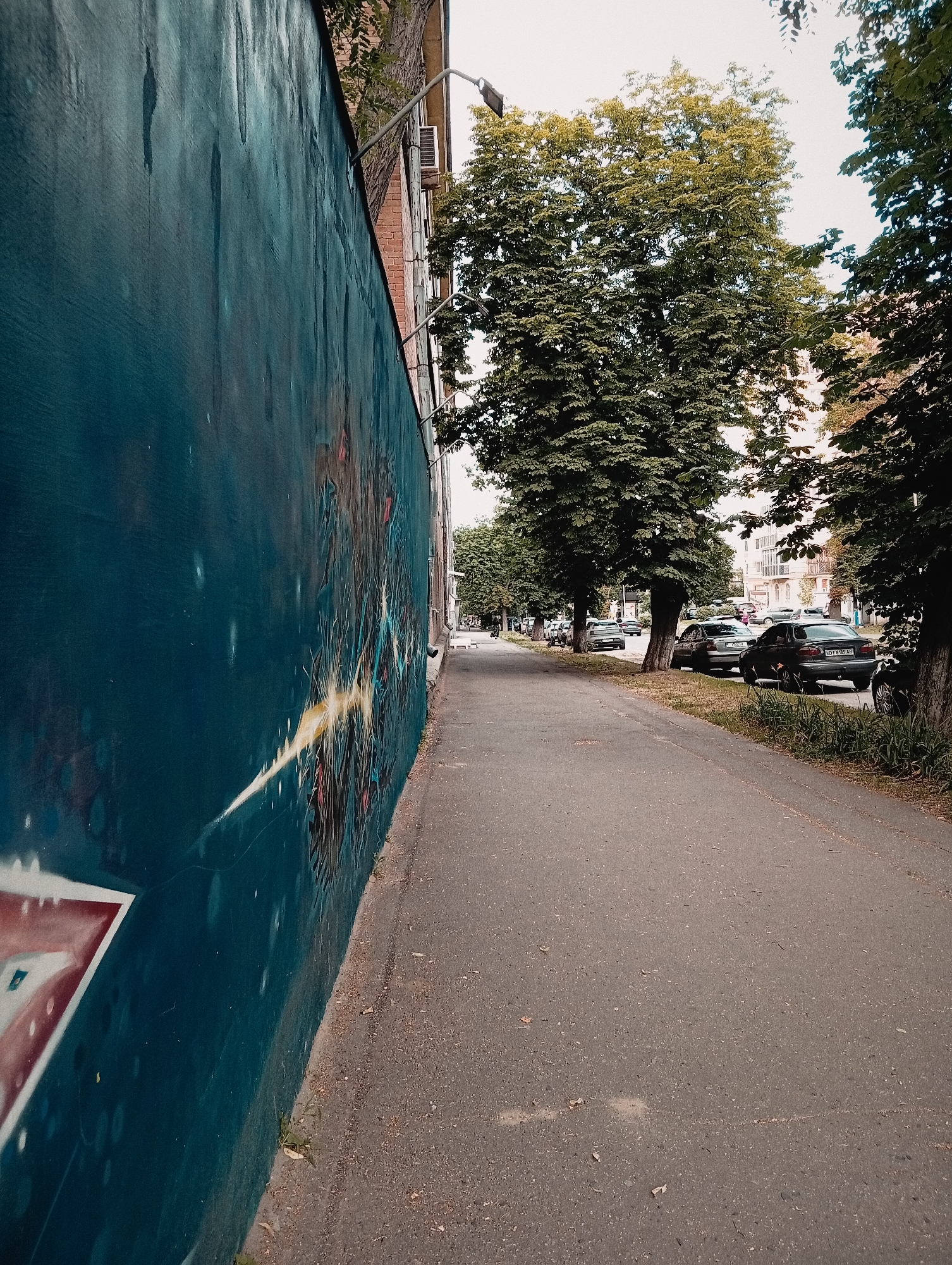 The image depicts a street scene with a large mural on a wall to the left, a sidewalk, and a street lined with parked cars and trees. The mural is abstract and colorful, with a prominent dark blue background and streaks of yellow, red, and white paint. The wall itself appears to be a building's exterior.
To the right of the mural is a paved sidewalk, which then transitions into a street. Several cars are parked along the side of the street, with more vehicles visible further down the road. The street is lined with mature trees that cast shadows on the sidewalk and road, suggesting it is daytime. The foliage of the trees is dense and green, indicating a warm season. The sky is overcast or the light is diffused, giving the scene a somewhat muted feel.
There is no clear activity or event taking place; it appears to be a typical urban street scene. Notable details include the graffiti on the wall, the variety of cars, and the consistent line of trees. No people are clearly visible in the frame. There is some text on a license plate of a dark-colored car, which appears to read "OT 695 AC". The overall impression is that of a quiet residential or commercial street, possibly in a European city, given the style of the cars and architecture in the background. The time of day is likely afternoon, given the lighting and shadows. The weather appears to be mild and possibly cloudy.