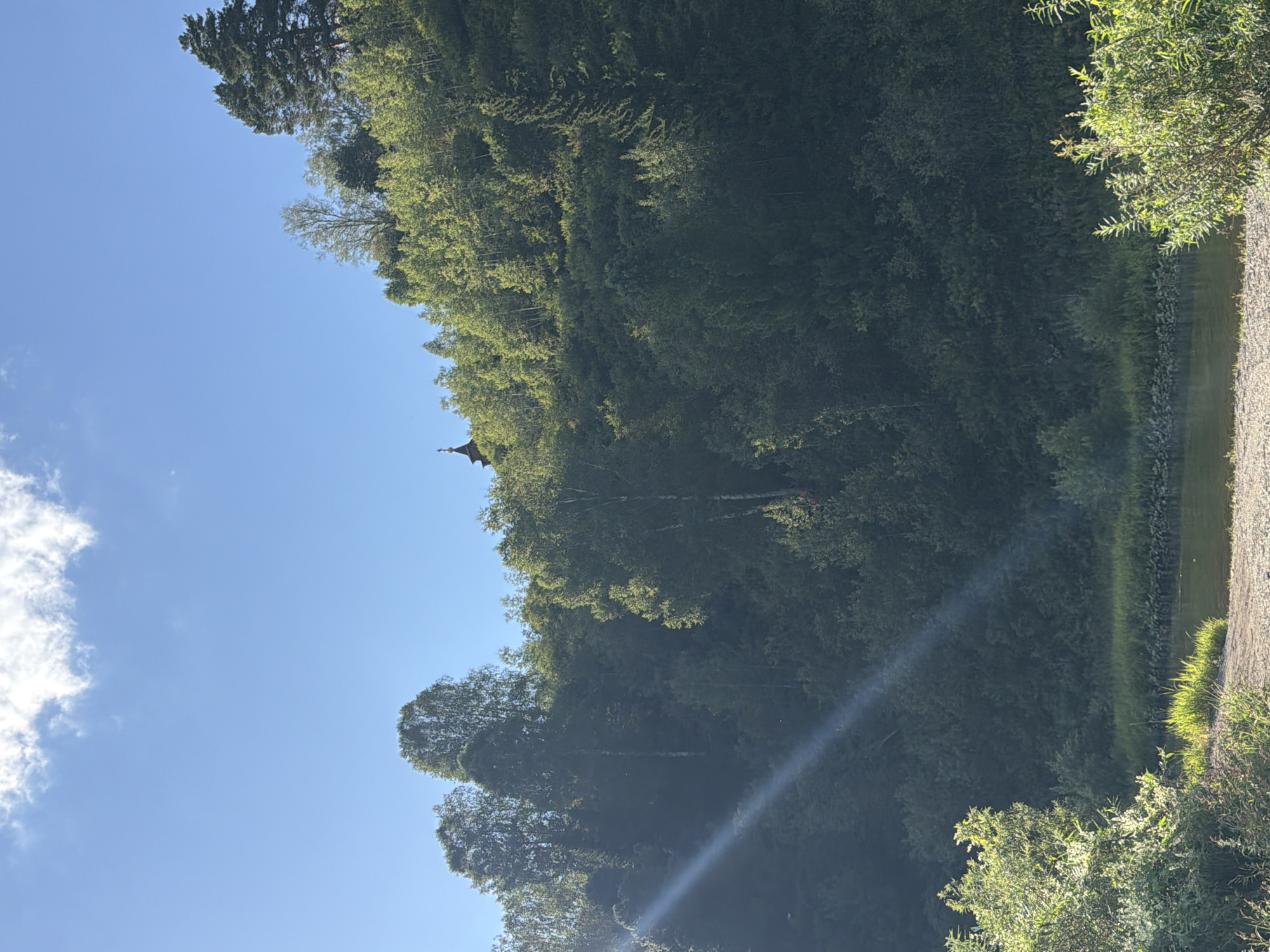 A sunlit slope covered with lush green trees rises from a calm river. A faint sunbeam cuts across the lower left quadrant of the image, illuminating some of the foliage. At the very top of the hill, a small, dark structure with a steeple, possibly a chapel, is visible. The sky above is a clear, bright blue with a few wispy white clouds. The foreground shows a rocky riverbank with some sparse vegetation. The scene is consistent with a natural landscape in a rural or wilderness setting.