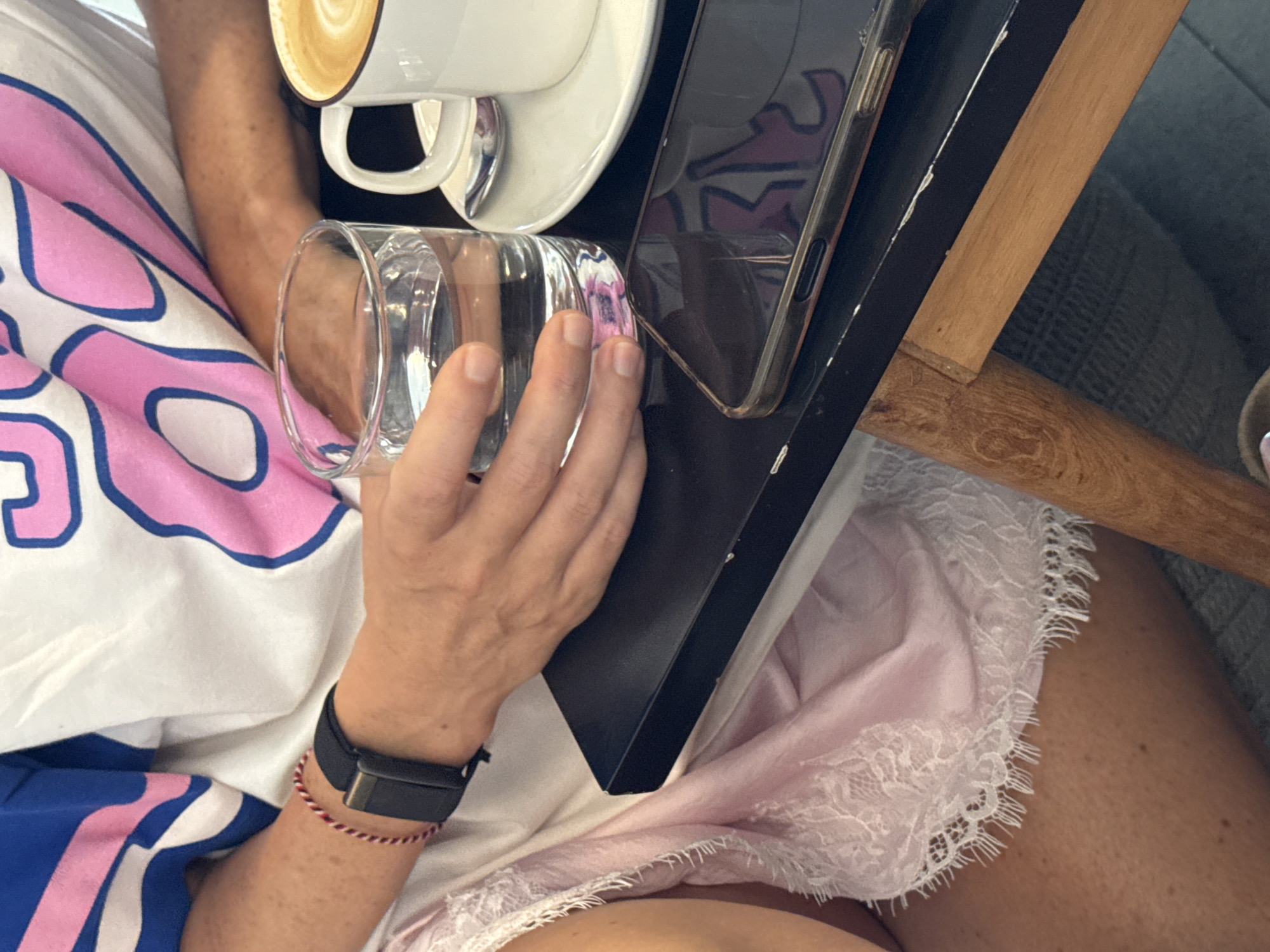A high-angle shot shows a person seated at a table in what appears to be a cafe or restaurant. The location is Kuta Selatan, Indonesia. The person is wearing a white shirt with pink and blue designs and a light pink garment with lace trim around the edge. They have a black smartwatch and a red string bracelet on their left wrist. The person is holding a glass in their right hand. On the black table is a cup of coffee, a saucer, and a smartphone.