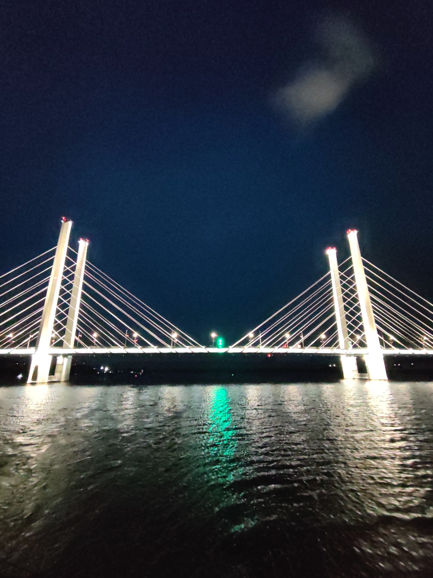 The image depicts a cable-stayed bridge at night. The bridge has two main towers, each illuminated with white light. Cables extend from the towers to the bridge deck. The bridge is lit with white lights along its length. There is a green light source at the center of the bridge. The sky is dark blue, with a single cloud visible. Below the bridge is water, reflecting the lights from the bridge.
This is the Oktyabrsky Bridge in Cherepovets, Russia.