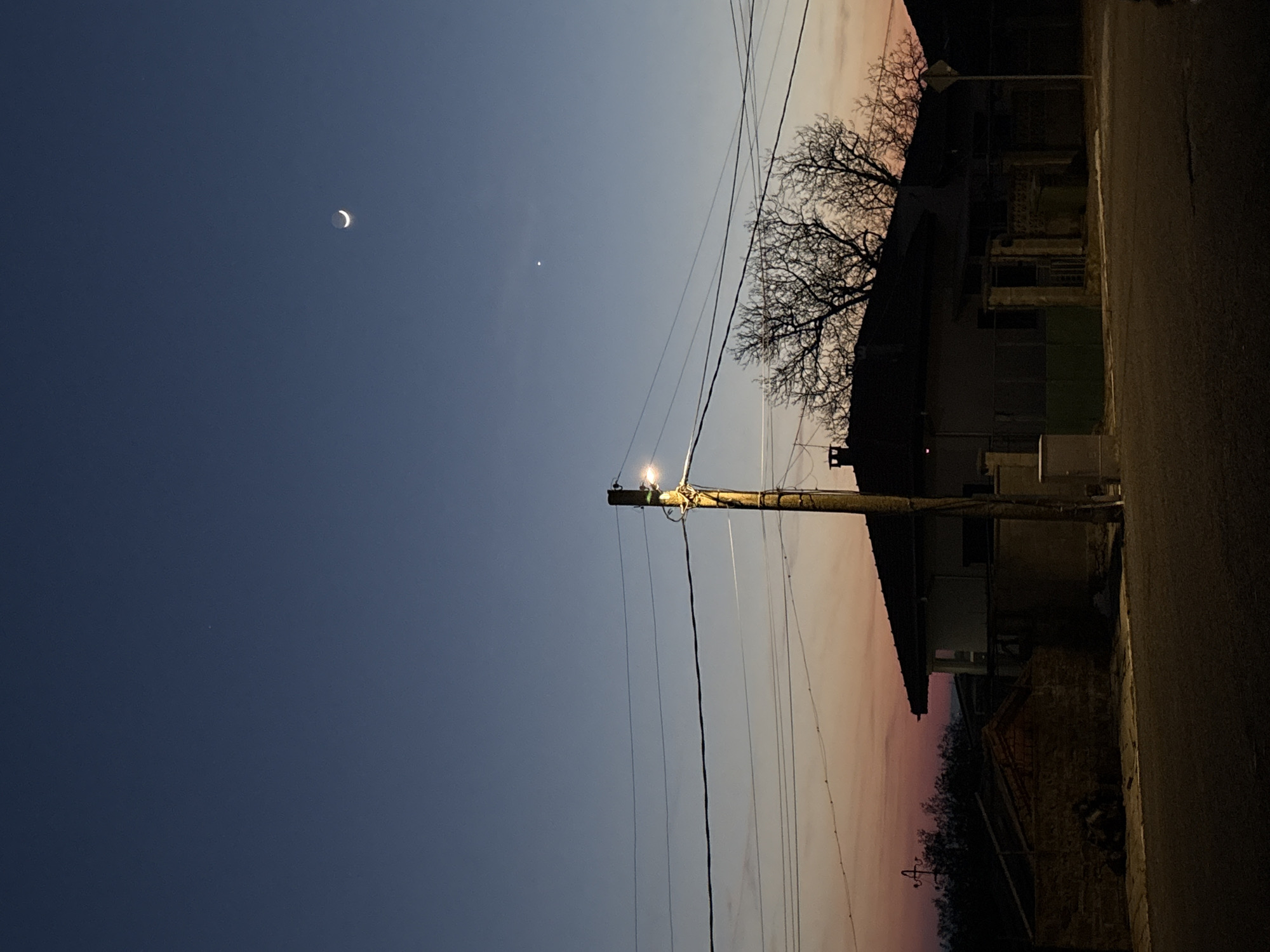 A low-angle nighttime shot in Goritsa, Bulgaria, shows a crescent moon and another point of light in the dark blue sky. A utility pole with a lit bulb is centered. Power lines extend from the pole. In the right side of the image is a dark street, a house and a bare tree silhouetted against the sky with a light pink and purple tinge.