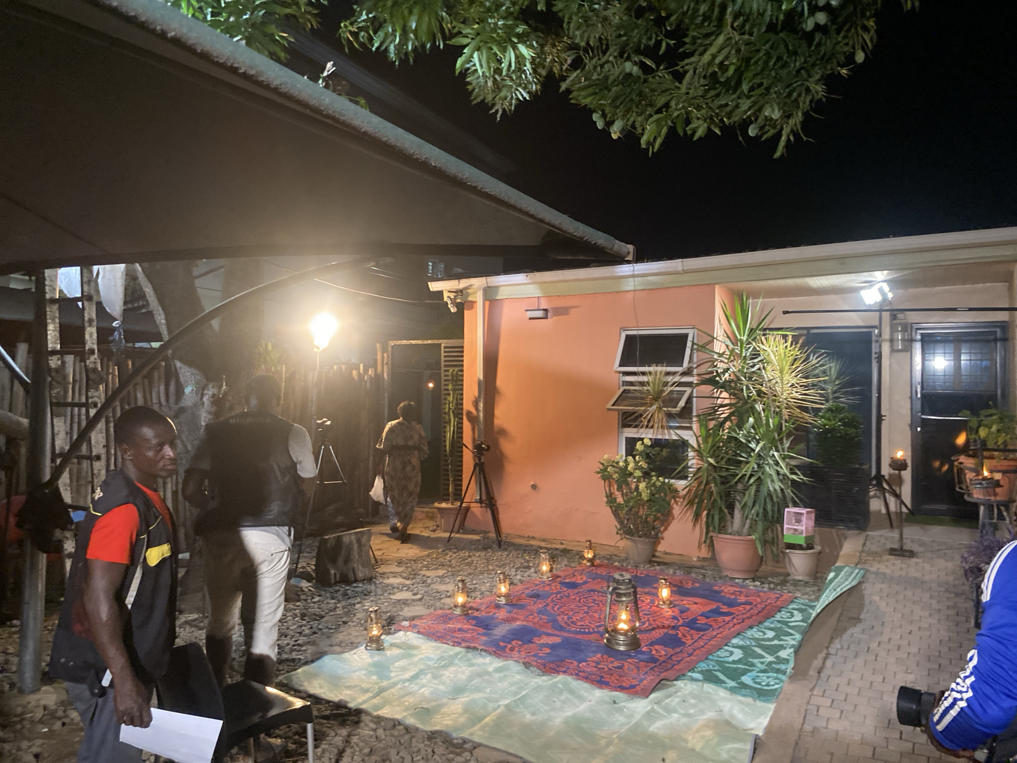 The image shows an outdoor film set at night. The scene is lit by several lanterns on a patterned rug placed on a light green sheet. A production light on a stand illuminates the area. Behind the rug is a one-story peach-colored building with a window and a black door to its right. Plants in pots are placed near the building. Two tripods with equipment are also visible.
On the left side of the frame, a man in a black vest and red shirt holds papers. Another man in a vest stands behind him, facing away from the camera. A woman in a patterned dress walks towards the building. On the right, part of a person wearing a blue jacket with white stripes is visible, holding camera equipment.
The location appears to be a backyard or courtyard, with a variety of foliage overhead. The image suggests a film production taking place at night, possibly in an urban or suburban area of Barden Yana, Nigeria. The presence of professional lighting and camera equipment implies a deliberate set-up for filming.
