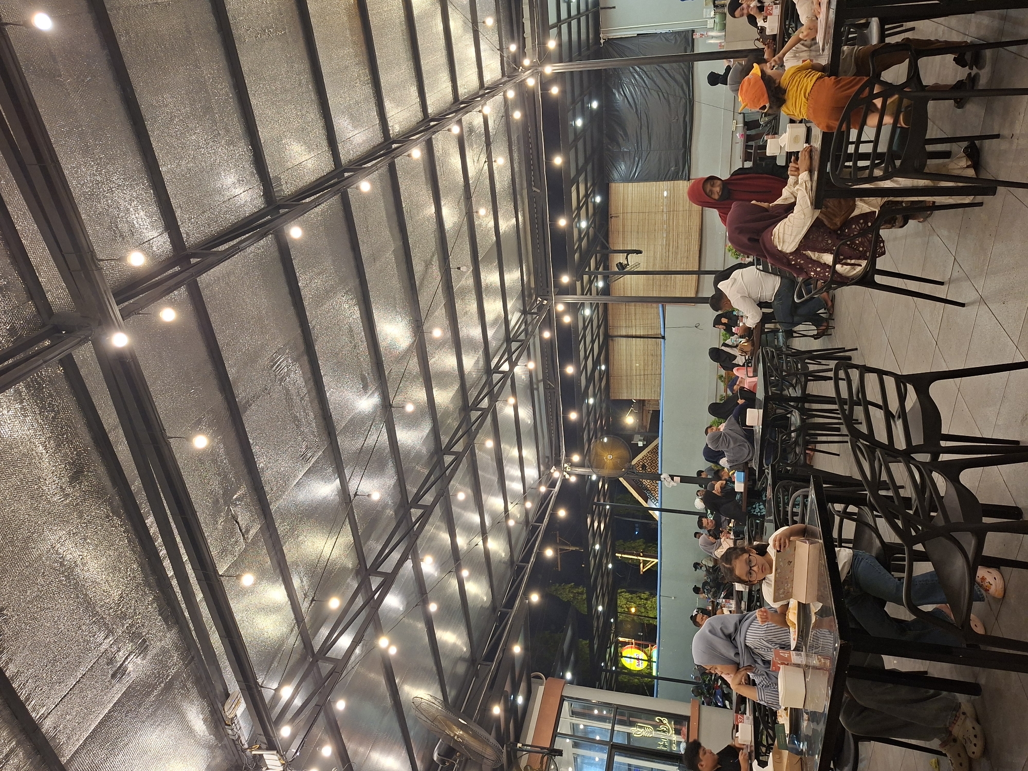 The image depicts a bustling indoor-outdoor restaurant in Lhokseumawe, Indonesia, filled with patrons at various tables. People are seated and engaged in dining, with some conversing. The restaurant features a high ceiling with exposed steel beams and a reflective lining, illuminated by strings of lights. The building opens to the outside with large windows revealing the night. A ceiling fan is visible near the window. The chairs are a uniform black.