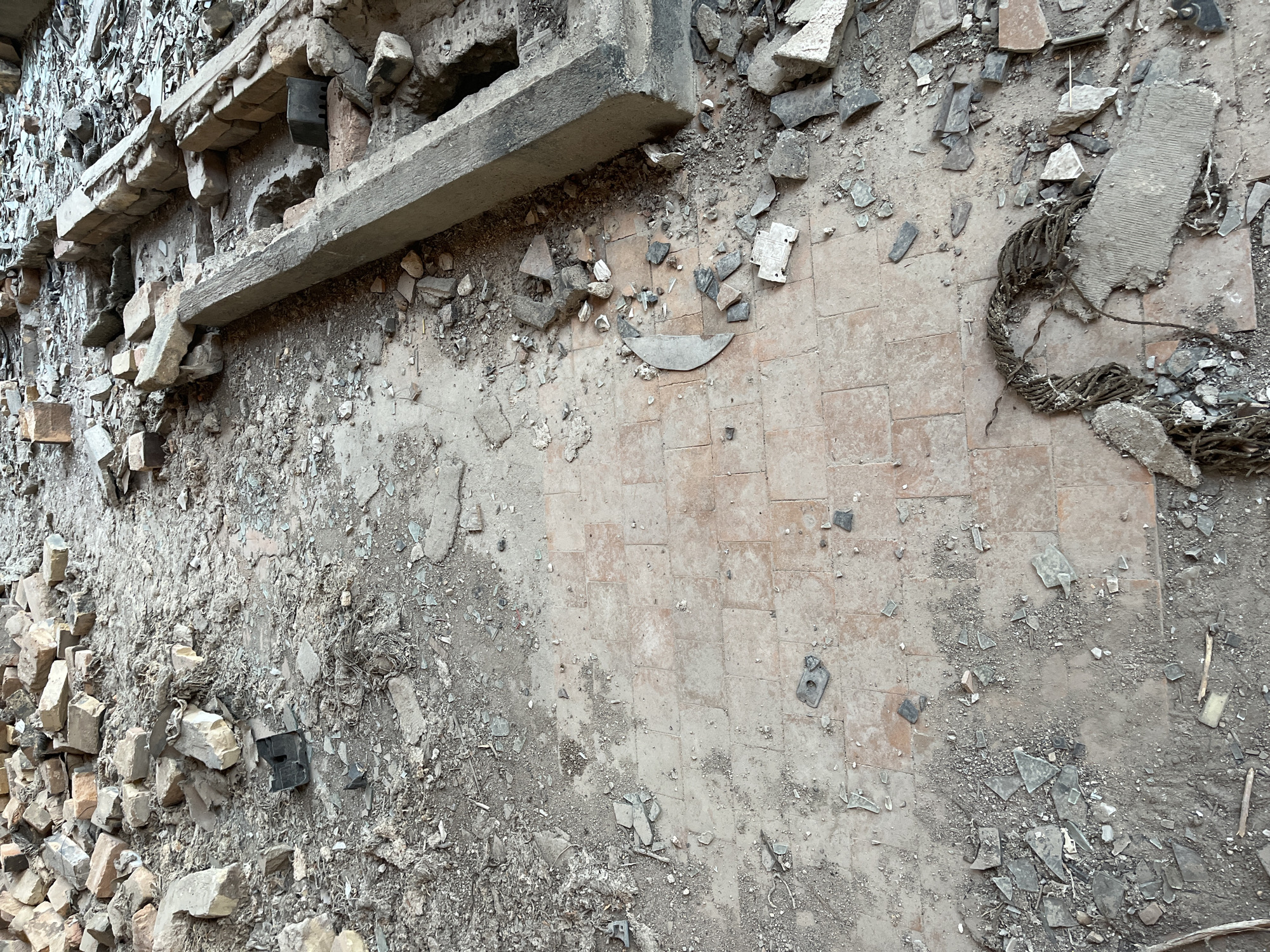 This image depicts a scene of destruction, likely following a demolition or natural disaster. The ground is covered in debris, including broken concrete, bricks, and what appears to be rebar or thick wire twisted into a coil. The terrain is uneven and dusty, with patches of what looks like tiled flooring peeking through the rubble.
In the upper left quadrant, a damaged concrete structure with exposed rebar and brickwork suggests the remnants of a building or wall. Small, dark, rectangular objects, possibly electrical components or fasteners, are scattered amongst the debris. A light-colored, flat concrete slab is prominently positioned in the center-right of the image, partially obscuring the tiled floor.
The lighting appears to be natural, suggesting daytime, but the weather is not discernible. The overall impression is one of disarray and abandonment, with no signs of human presence or activity. The location context indicates the scene is in Қарғалы, Kazakhstan, but there are no specific visual cues within the image to confirm this. No text is visible in the image.