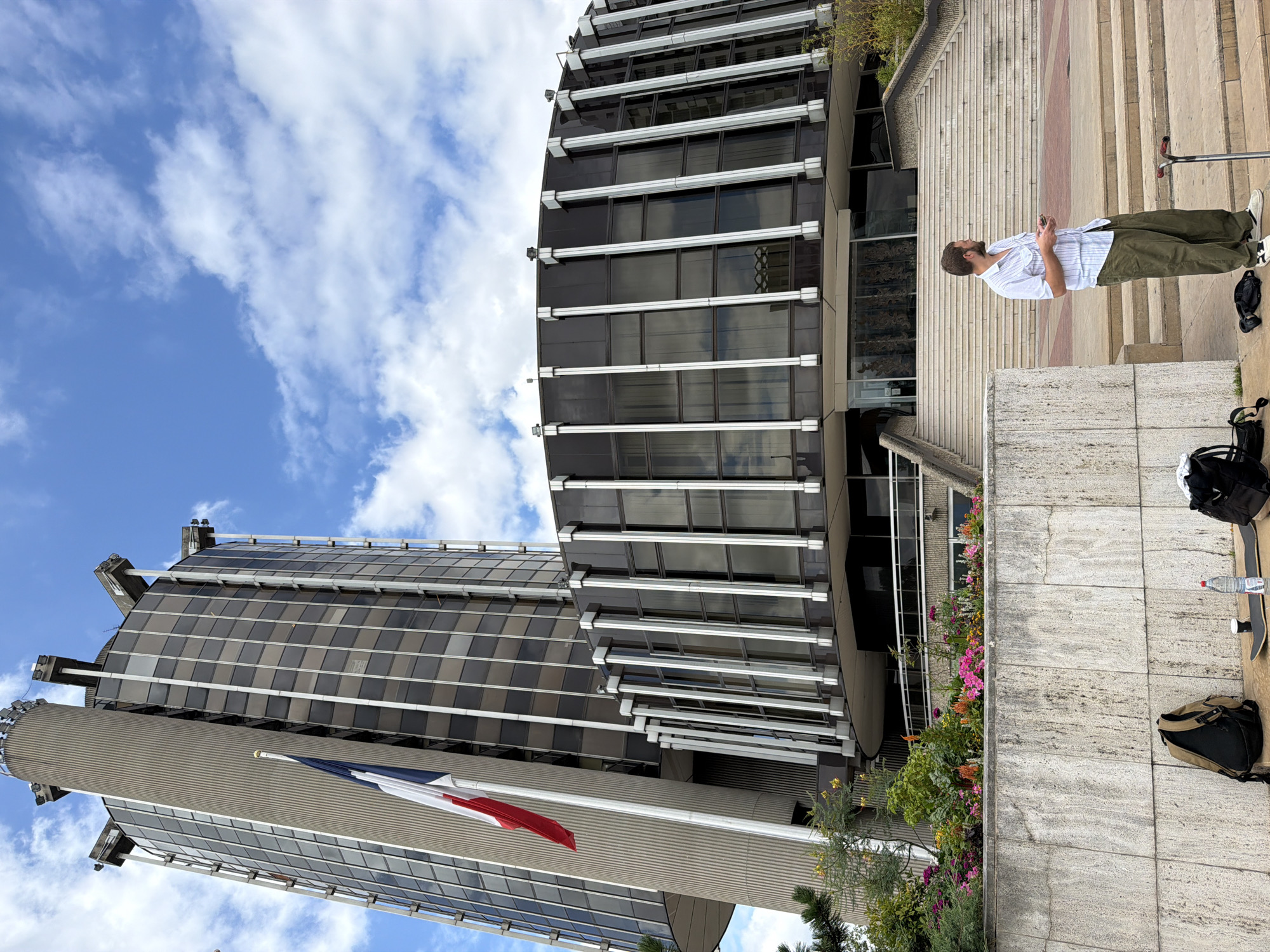 A man wearing a striped short-sleeved shirt and olive green pants stands on the steps of a building, holding a mobile phone. In front of him, a skateboard and a scooter are placed on the ground. Backpacks and a water bottle are also visible near the man. The building is a multi-story structure with a rounded glass facade. A French flag waves from a flagpole on the left side of the building. The sky is partly cloudy. This is the Hôtel du Département in Créteil, France.