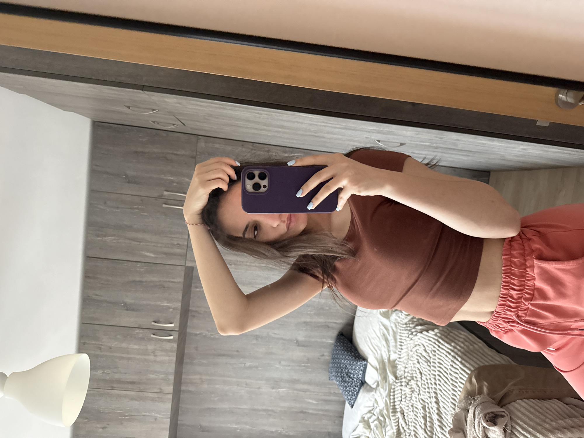 A woman is taking a selfie in what appears to be an indoor setting. She is holding a purple cell phone up to her face, obscuring some of her facial features. The woman is wearing a brown short-sleeved crop top and coral-colored sweatpants. She is touching her hair with her free hand. Behind her, there is a bed with patterned bedding and pillows. The background also includes a gray closet or cabinet and a lamp. There is no definitive evidence in the image to confirm the location is Plovdiv, Bulgaria. The image is taken in a mirror, resulting in a mirrored perspective of the environment.