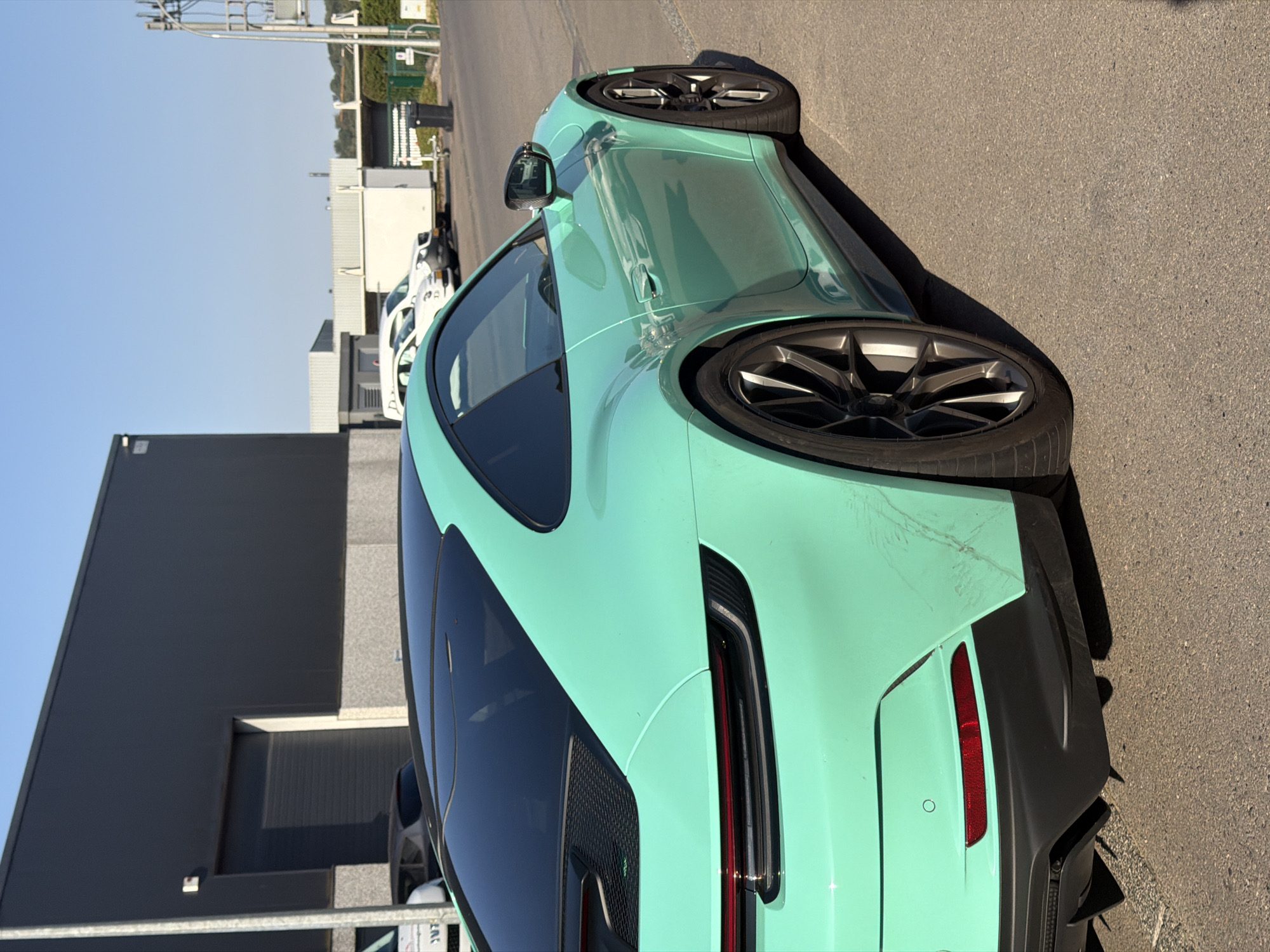 The image shows the rear-right side of a turquoise Porsche 911 GT3 parked on an asphalt surface. The car is angled slightly away from the viewer, with its rear bumper, tail light, and one wheel clearly visible. The car has a black roof and black accents. In the background, a large, dark gray industrial building with a closed roll-up door is situated to the left. To the right and further back, there are several white vehicles, including what appears to be a small trailer and a black trash bin. Beyond these, a line of green bushes and trees is visible, with a bright blue sky above. The scene appears to be in an industrial or business park area, likely in Leudelange, Luxembourg.